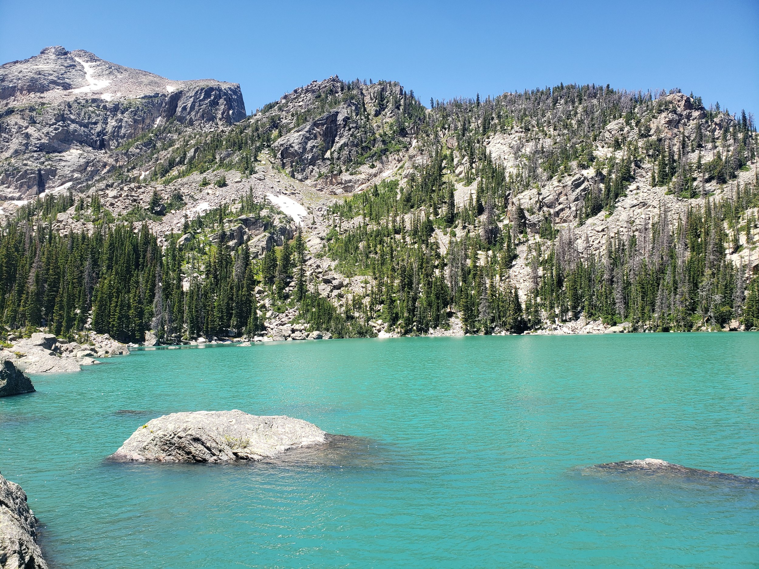 A turquoise lake with rocks along the shore, Lake Haiyaha, surrounded by evergreen trees and tall rocky mountain cliffs under a clear blue sky. Photo taken by Femme Trek on a women-only Lake Haiyaha guided hiking tour with Femme Trek