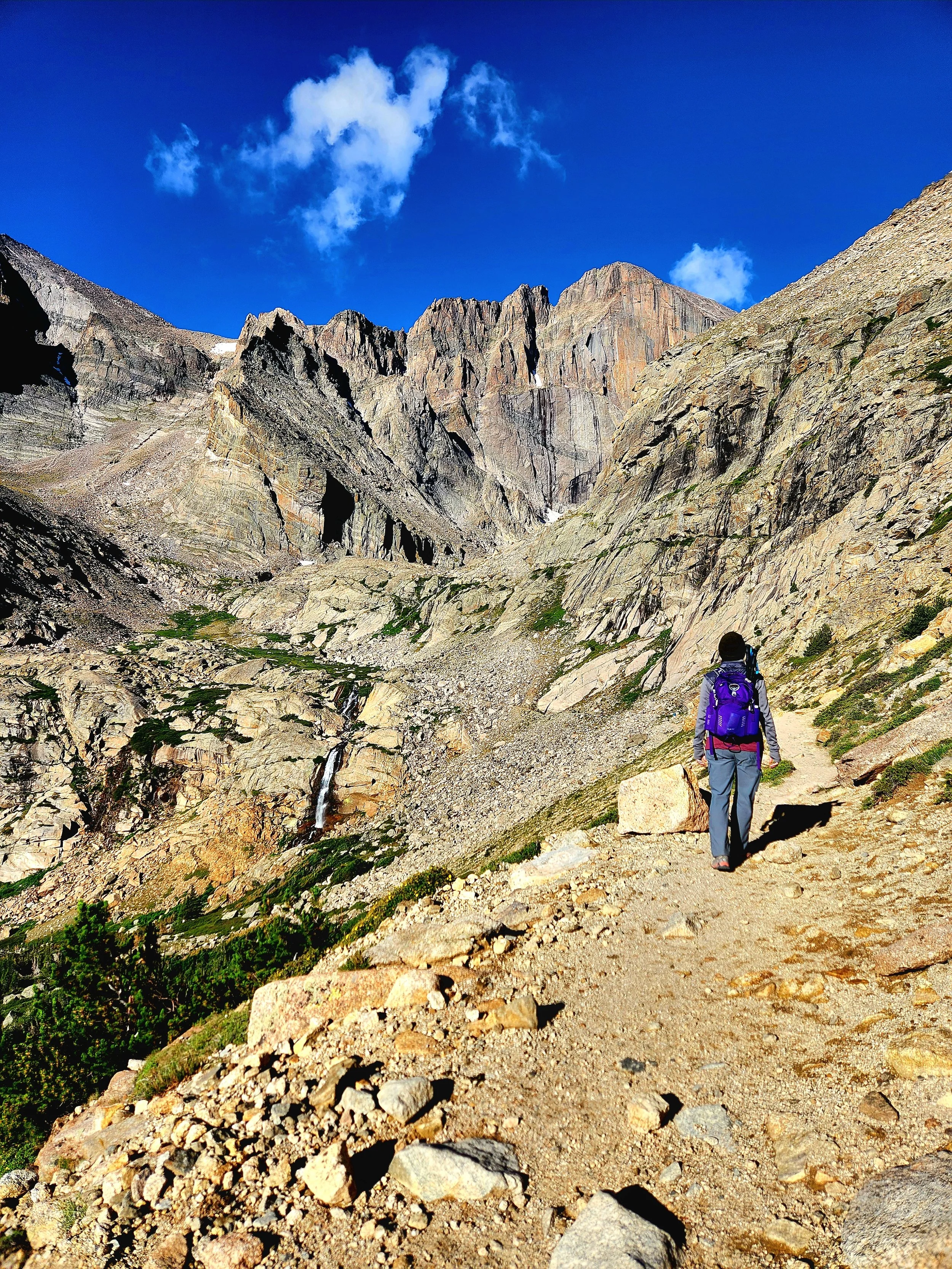 A woman with a purple backpack hiking on a rocky mountain trail with rugged mountain peaks and a partly cloudy blue sky in the background. Photo taken on a women-only guided hiking tour to Chasm Lake in Rocky Mountain National Park with Femme Trek.