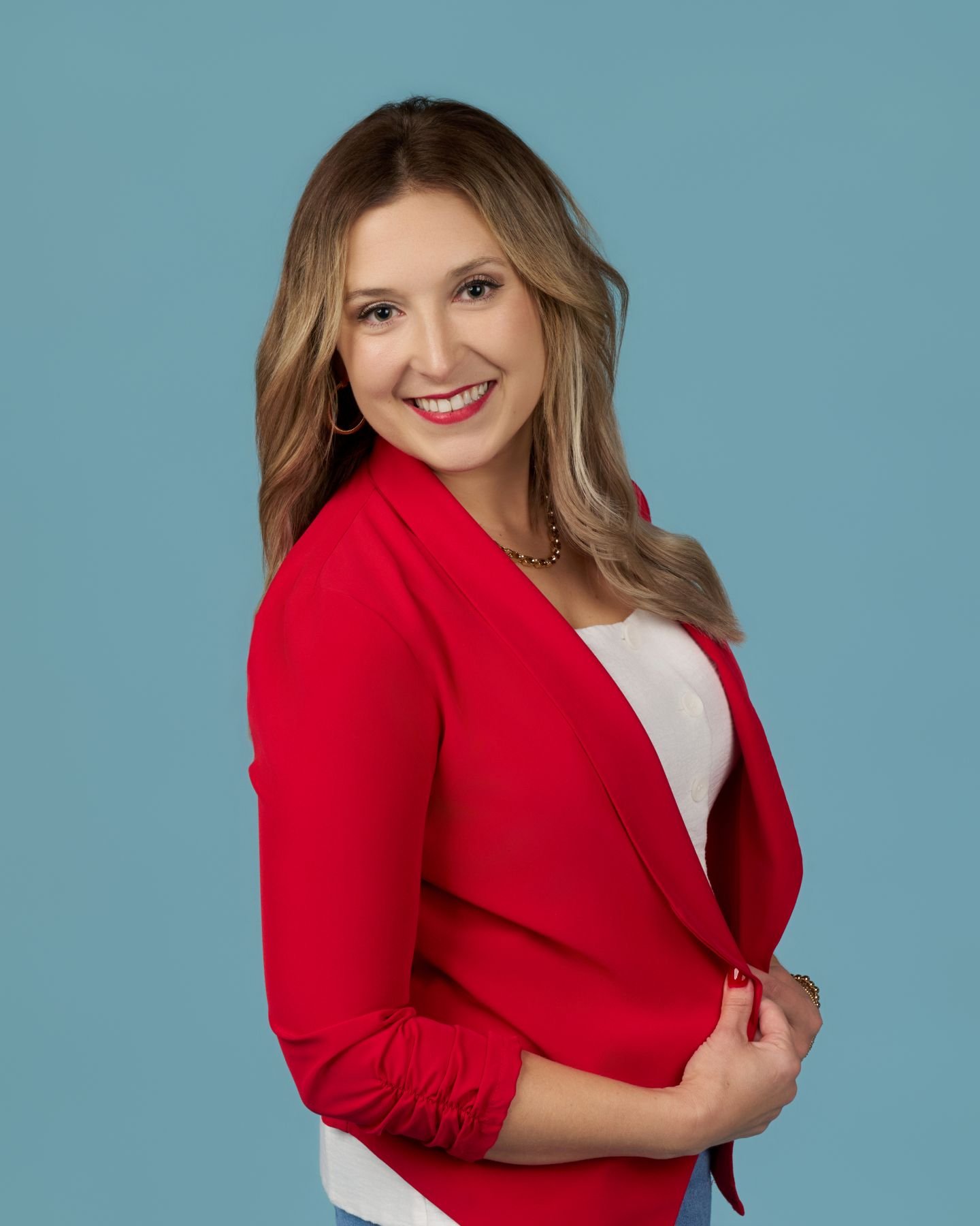 A woman with shoulder-length blonde hair smiling, wearing a red blazer over a white top, standing against a light blue background.