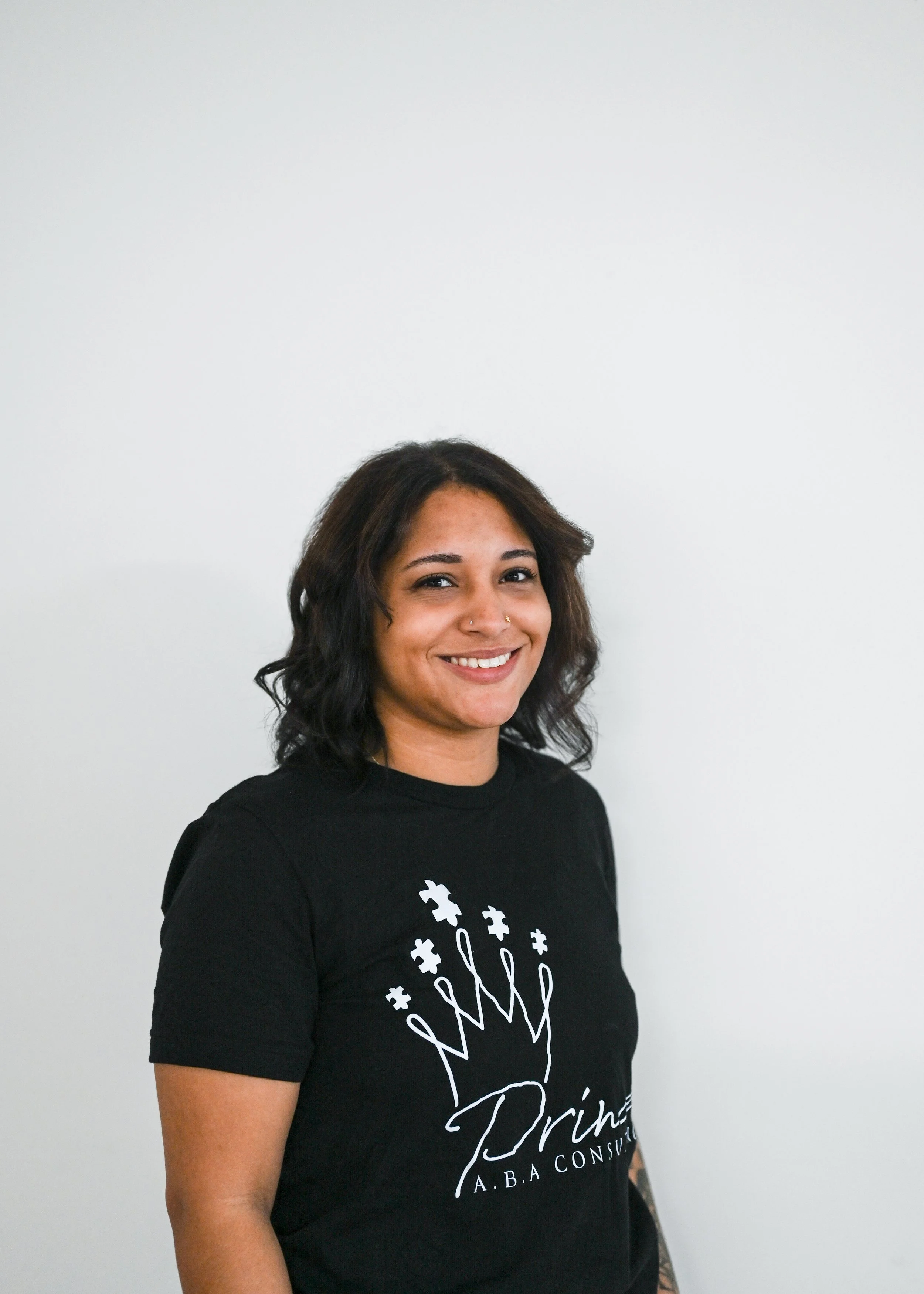 A smiling woman with dark brown, shoulder-length wavy hair, wearing a black T-shirt with a white floral crown and text design, standing against a plain white background.