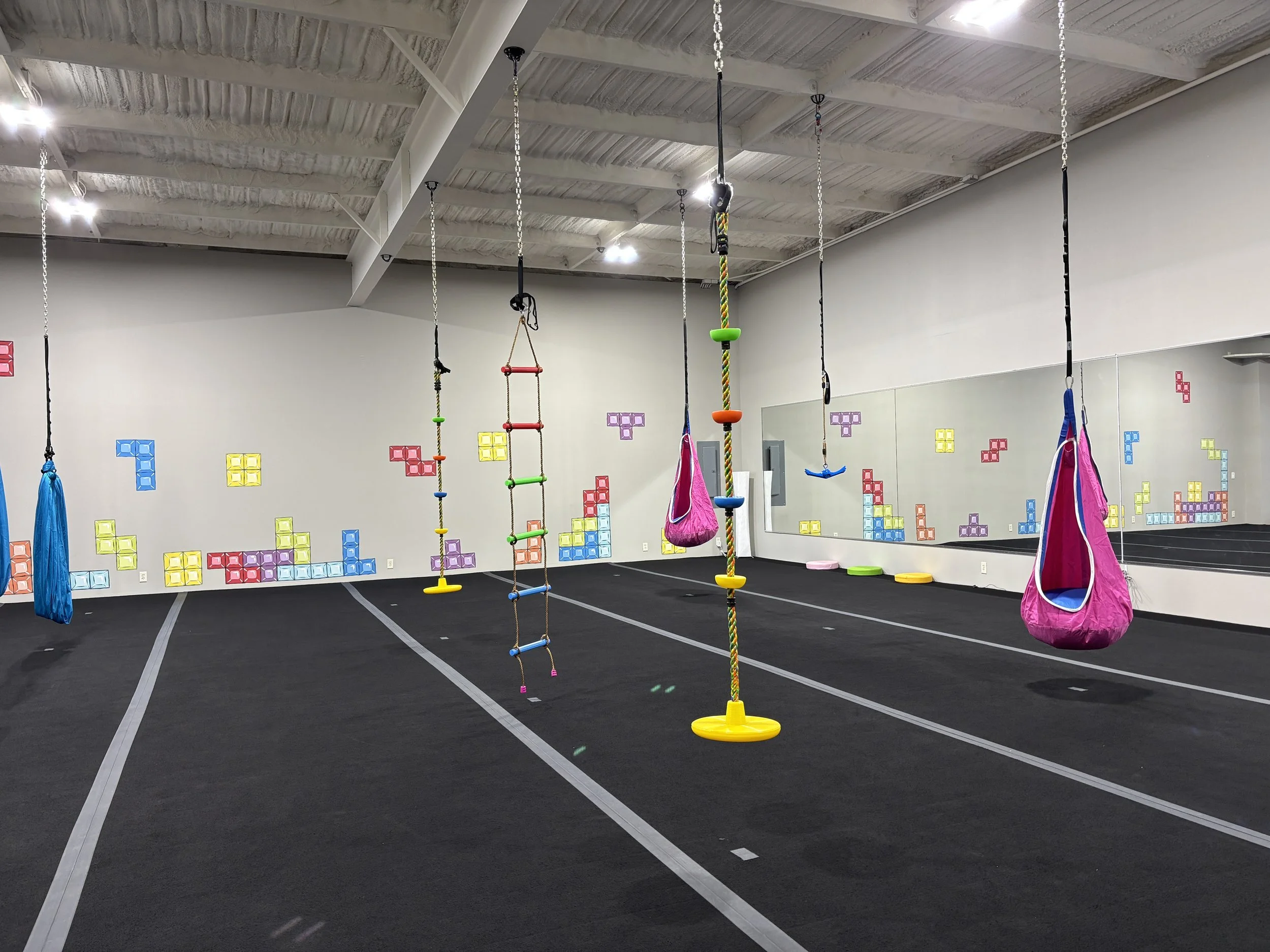 Large gym room with tetris wall mural.  Hamocks, ladders, and swings hang from the ceiling and a wall mirror.  