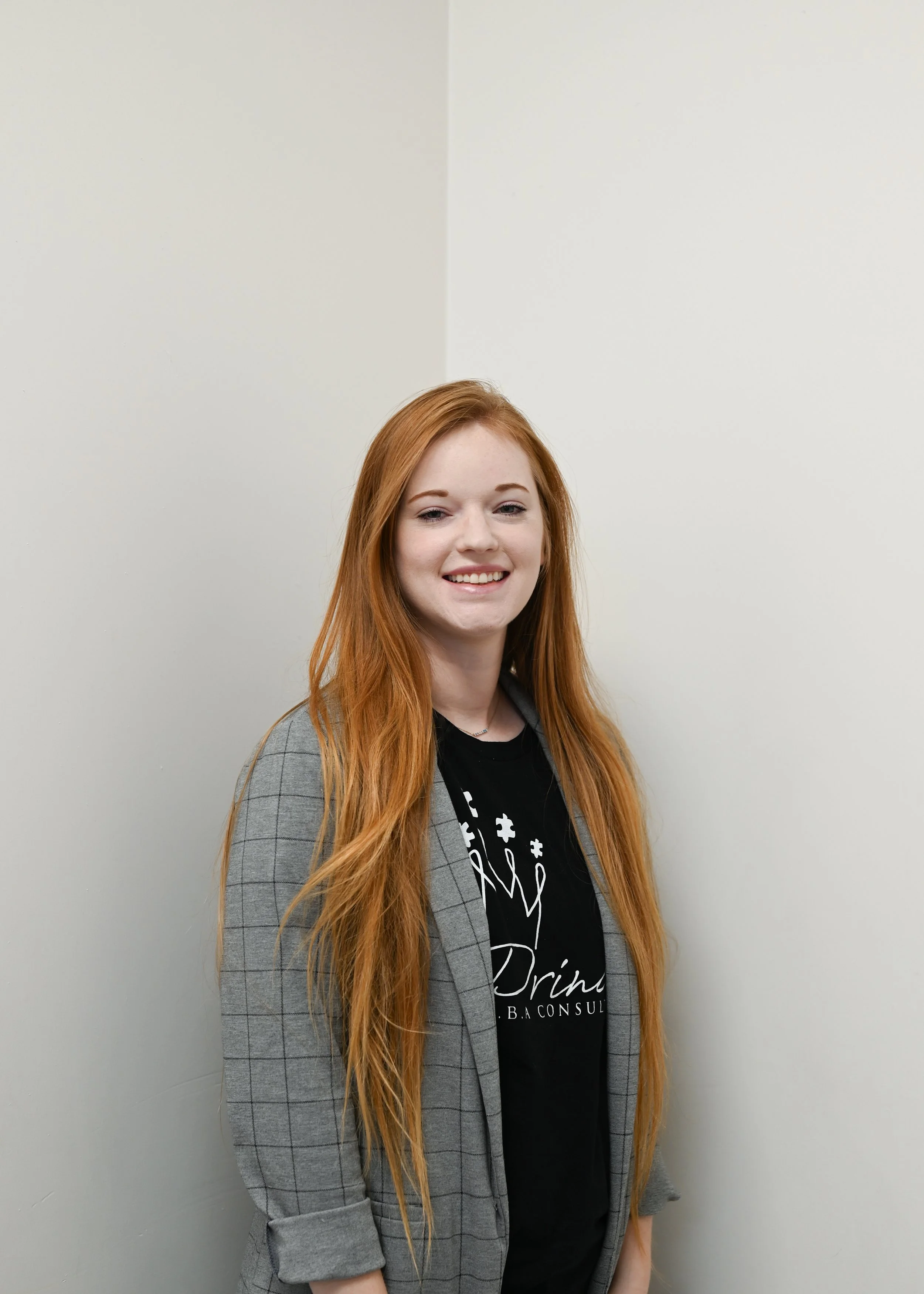 A woman with long red hair wearing a gray checkered blazer and a black T-shirt with white text, standing against a plain off-white wall.