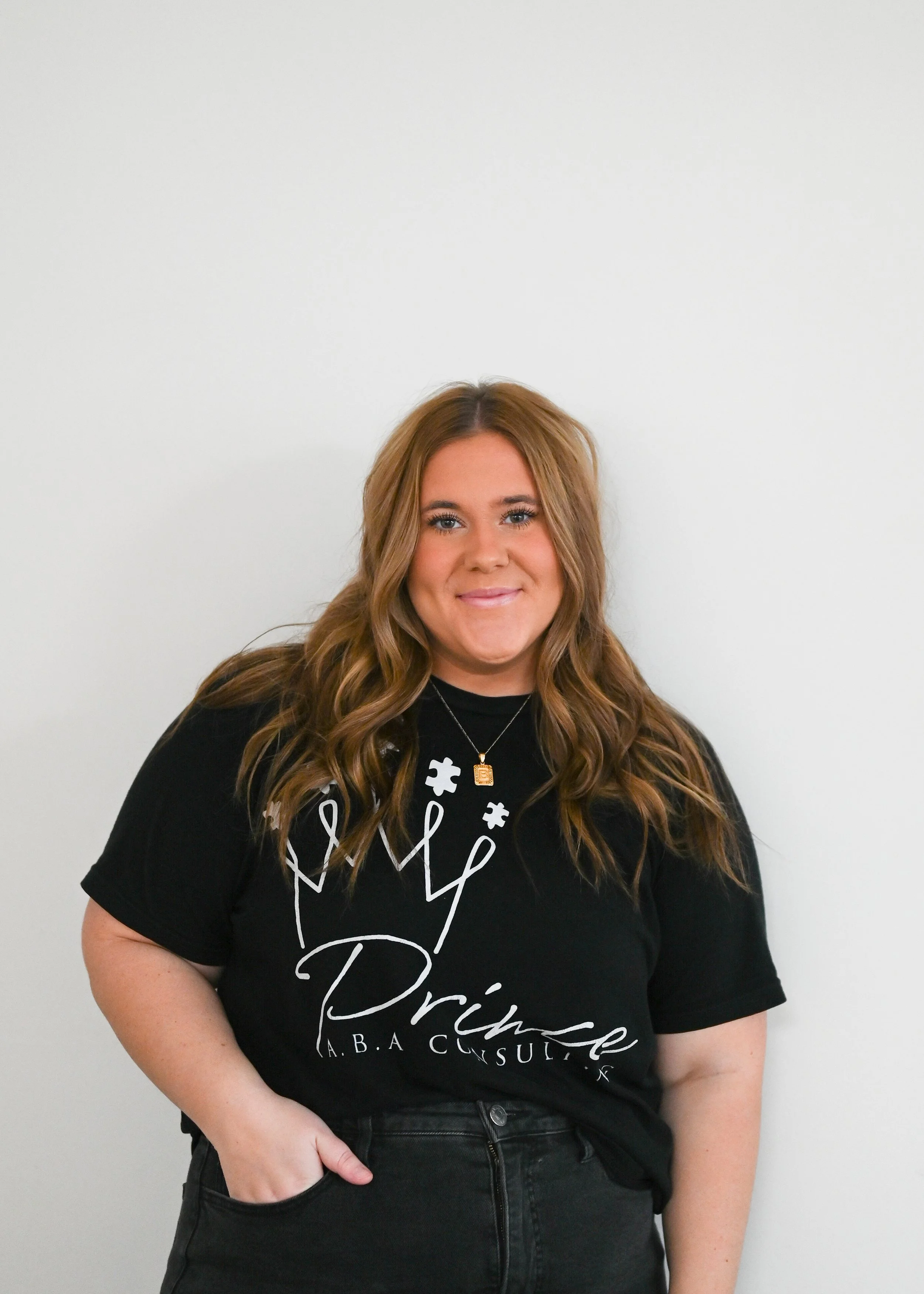 A woman with long wavy brown hair wearing a black t-shirt with white text and graphics, standing against a plain white wall.