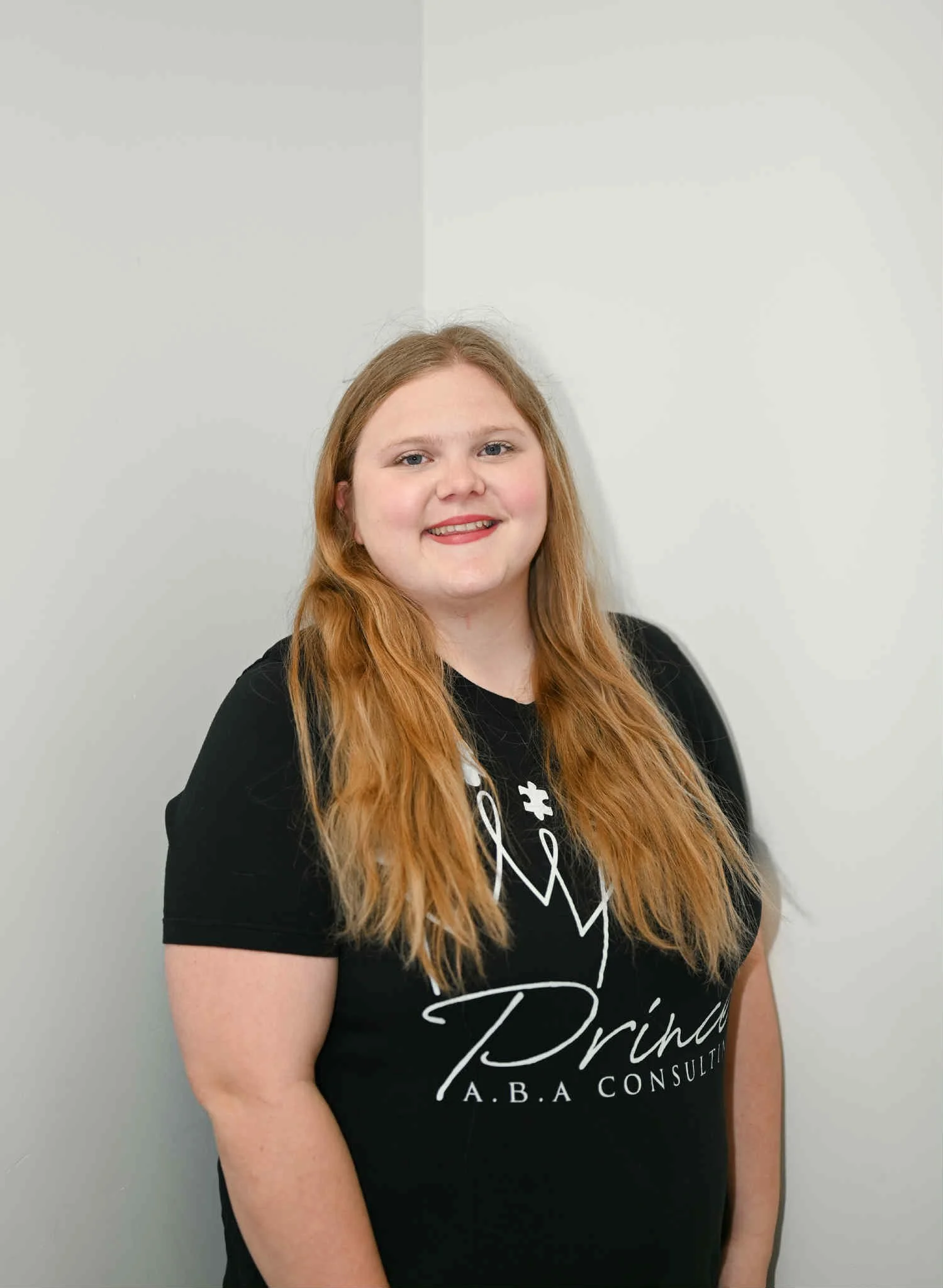 A woman with long, red hair smiling while standing against a plain white wall, wearing a black T-shirt that reads 'Princess A.B.A Consulting' with a stethoscope graphic.