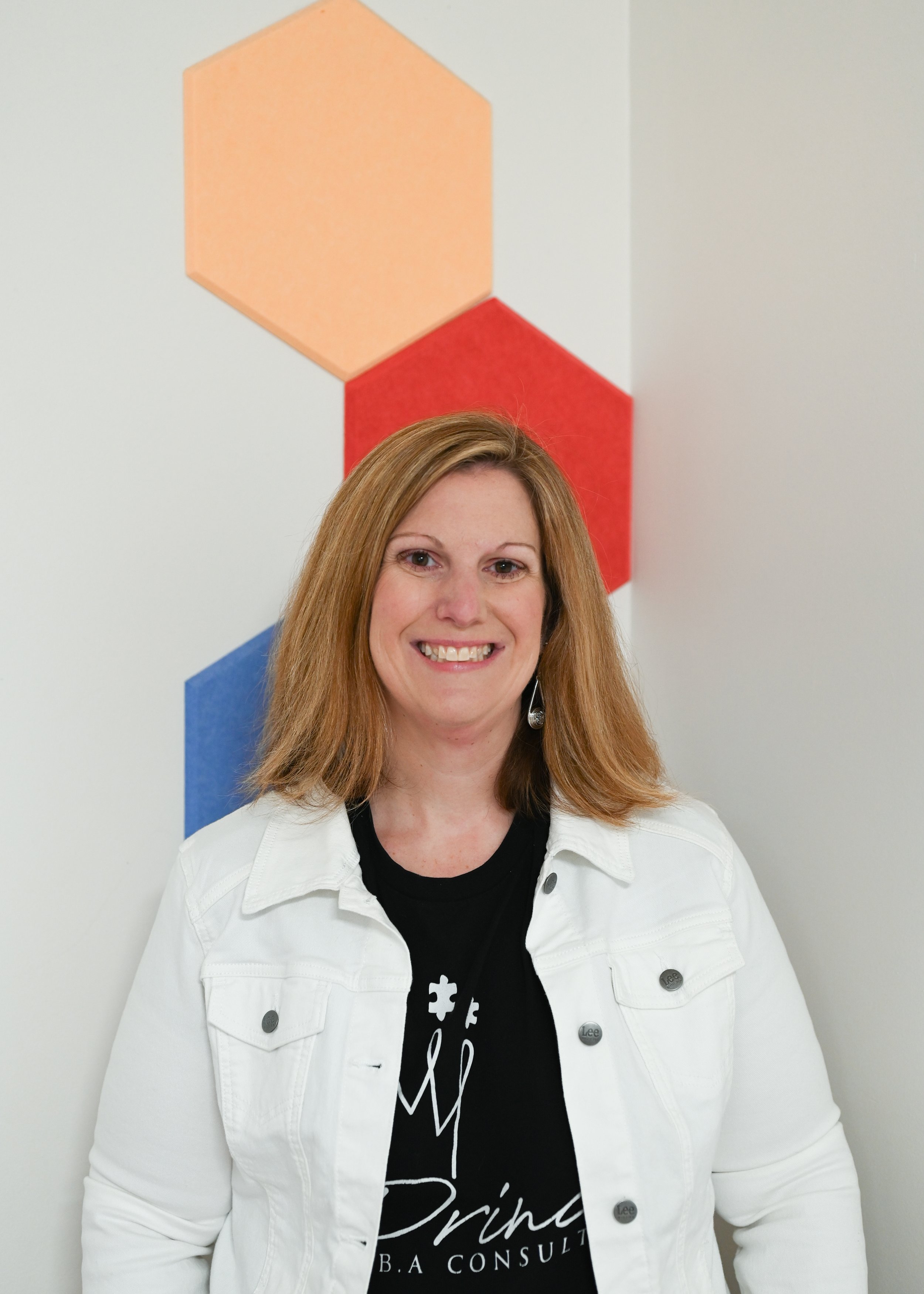 A woman with shoulder-length red hair smiling, wearing a white denim jacket and a black t-shirt with white text, standing in front of a wall with geometric colored shapes.