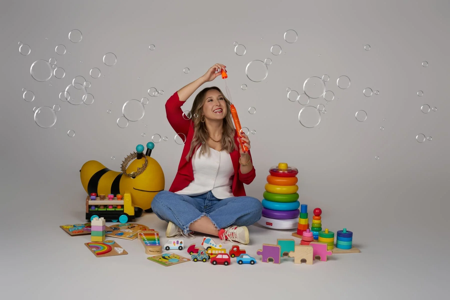 A young woman sitting cross-legged on the floor, smiling, holding a bubble wand with bubbles floating around, surrounded by colorful toys including a bee-shaped toy, stacking rings, toy cars, puzzle pieces, and wooden blocks, with a plain light gray background.