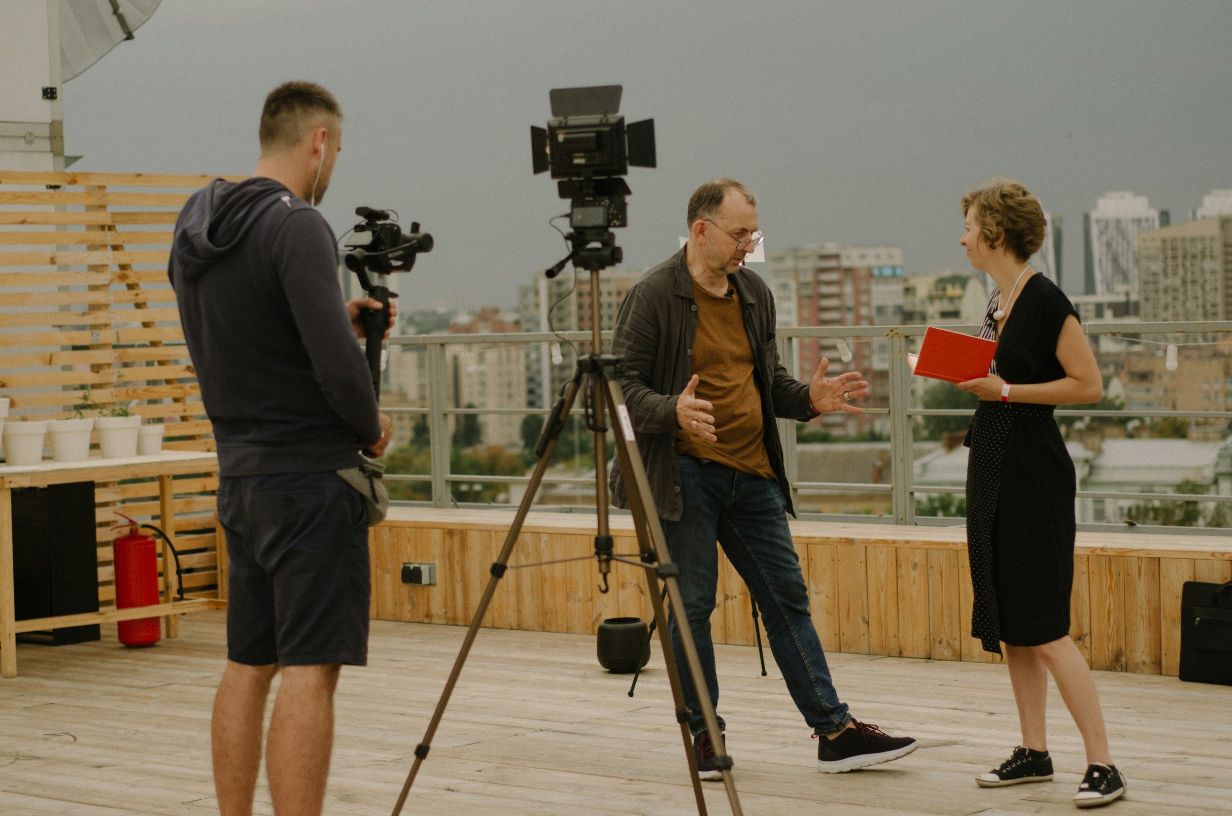 Three people on a rooftop, one with a camera, engaged in filming or interviewing, with city buildings in the background and cloudy sky overhead.