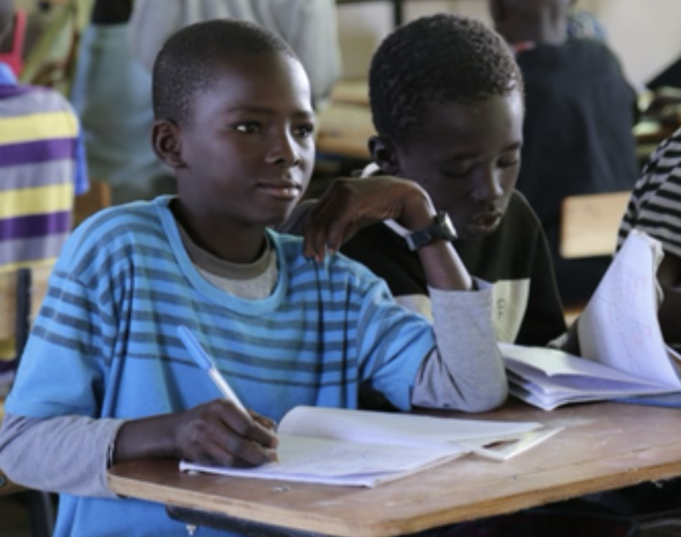Two boys sitting at a desk in a classroom, studying with notebooks and pens.