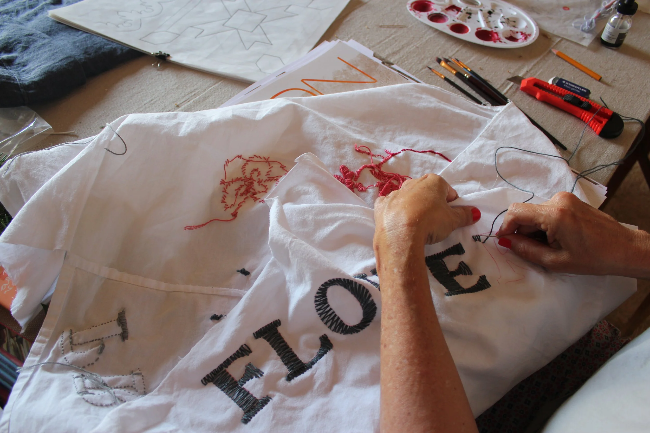 Person embroidering on white fabric with black letters and red threads, surrounded by sewing tools.