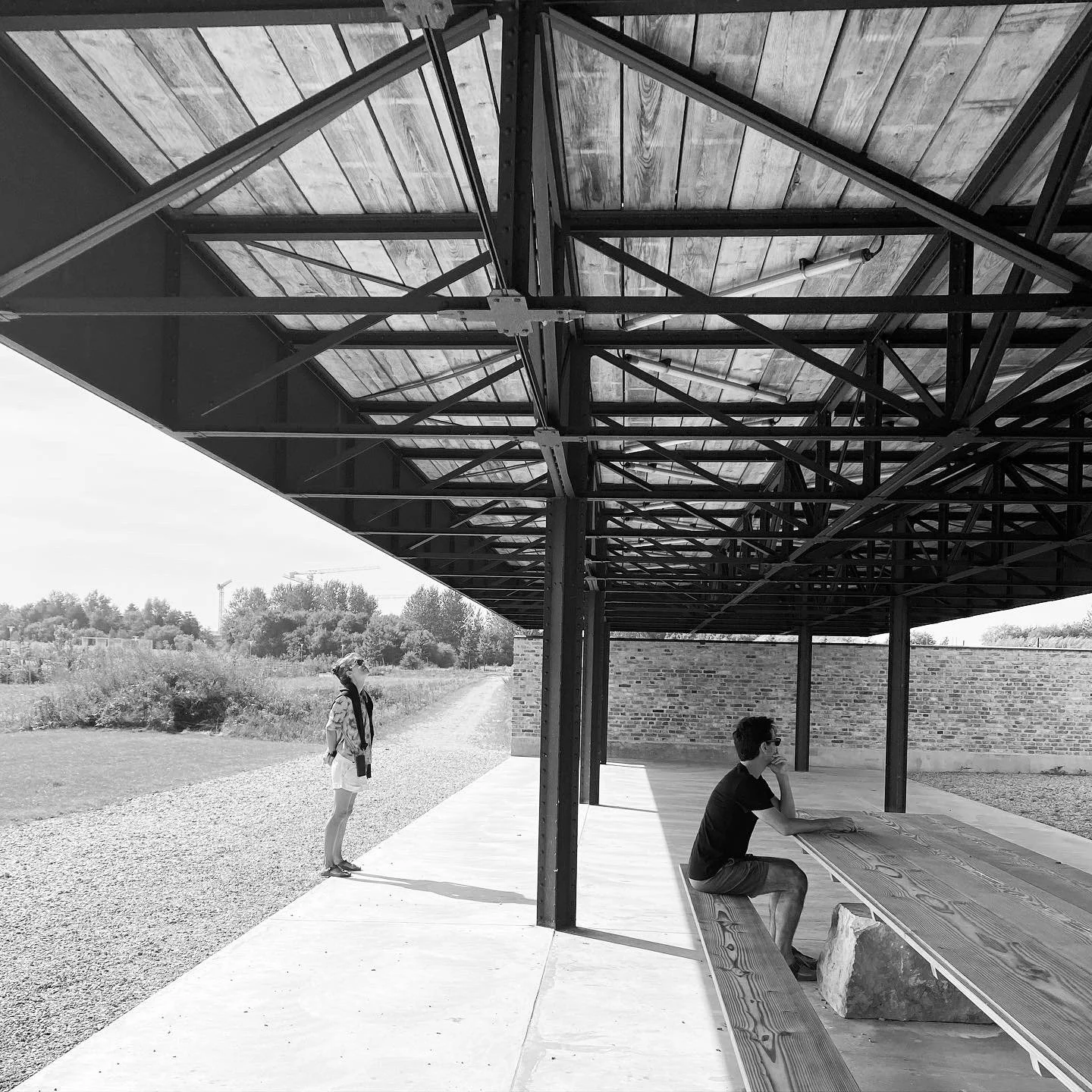 Black and white photo of two people under a large metal and wood structure with a brick wall, long wooden table, and benches outside.