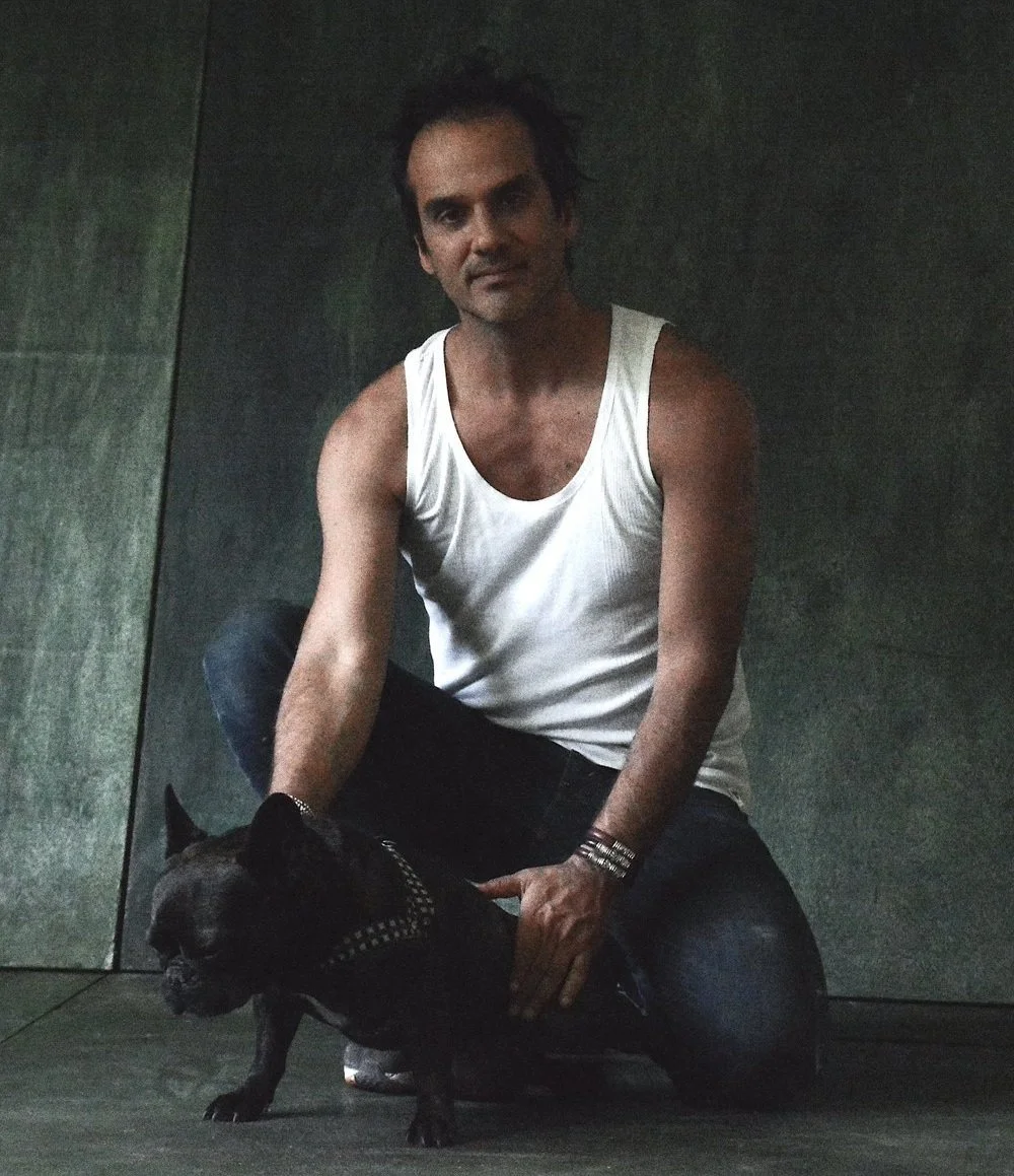 Man kneeling next to a black French Bulldog with a studded harness in a dimly lit setting.