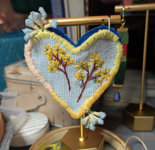 A decorative embroidered heart with yellow flowers hangs on a gold stand.