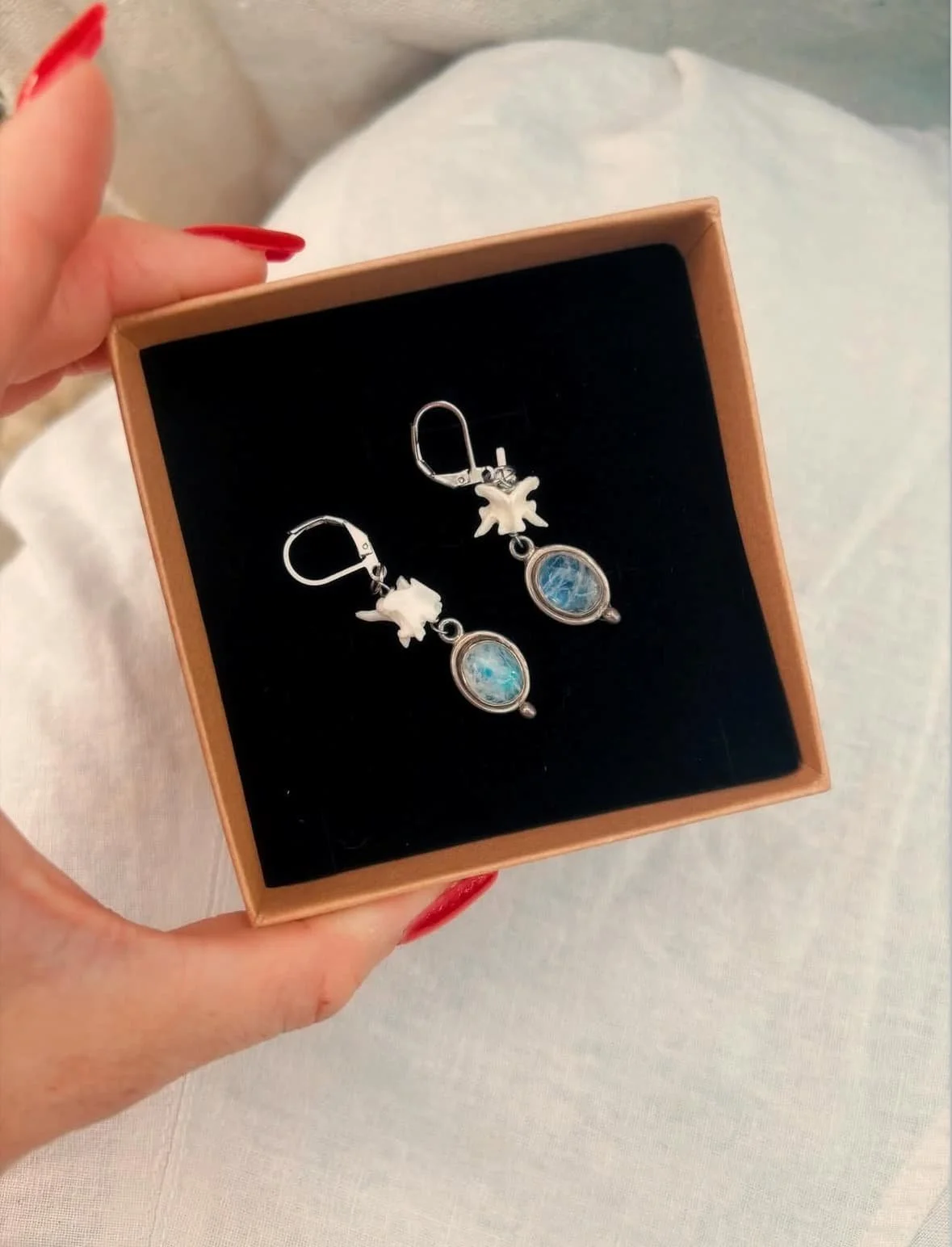 A pair of earrings and a pair of earrings in a box with a black interior. The earrings have oval blue stones and small white leaf-shaped charms.