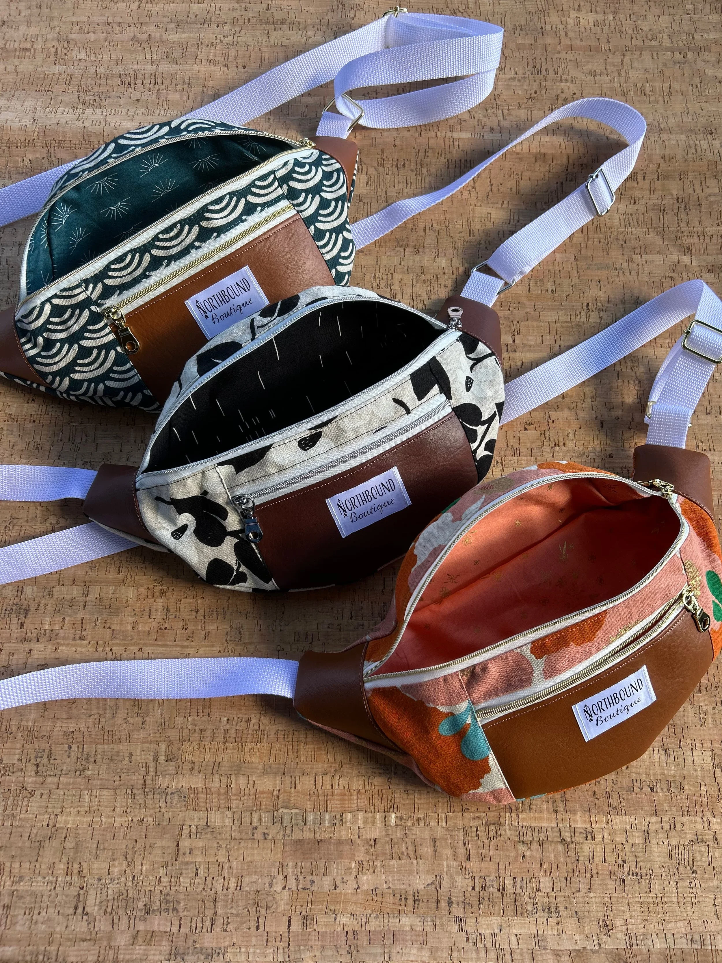 Three small fabric crossbody bags with white straps and metal buckles on a wooden surface. Each bag has a different pattern: one with black and white abstract floral, another with black and white animal print, and the third with a multicolored floral design. All bags have a label reading "NORTHBOUND Boutique."