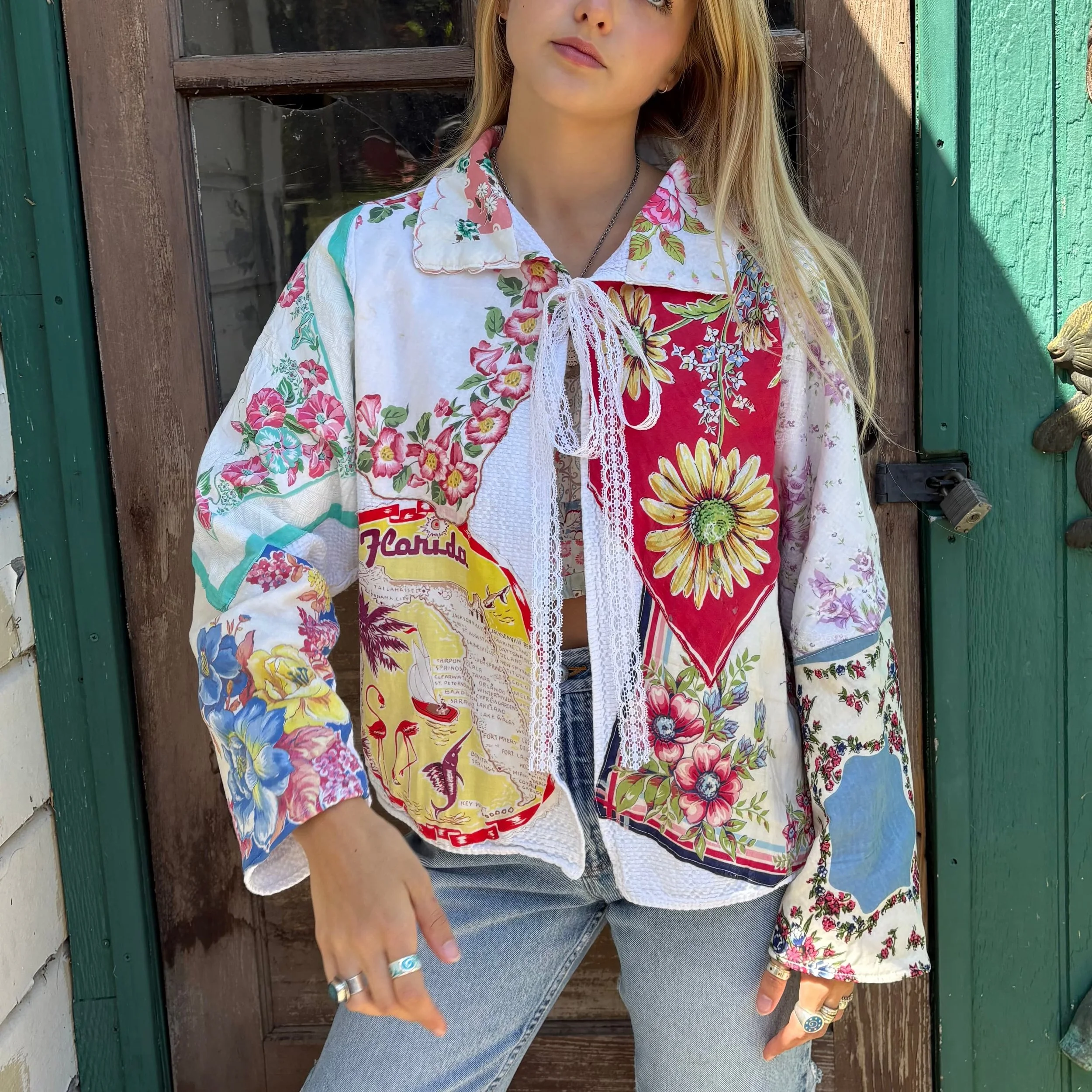 A young woman with long blonde hair stands outdoors near a wooden door. She is wearing a floral jacket over a white lace top, paired with blue jeans. She has rings on her fingers and wears a black necklace. She is pointing at her jeans with her left hand.
