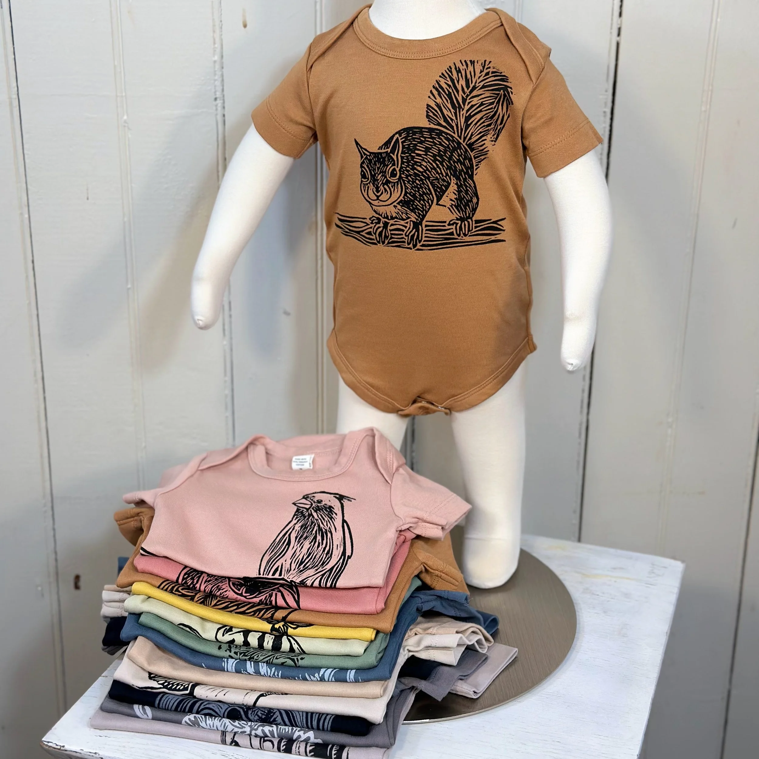 Stacked folded t-shirts on a wooden table, with one pink shirt featuring a black illustration of a bird.