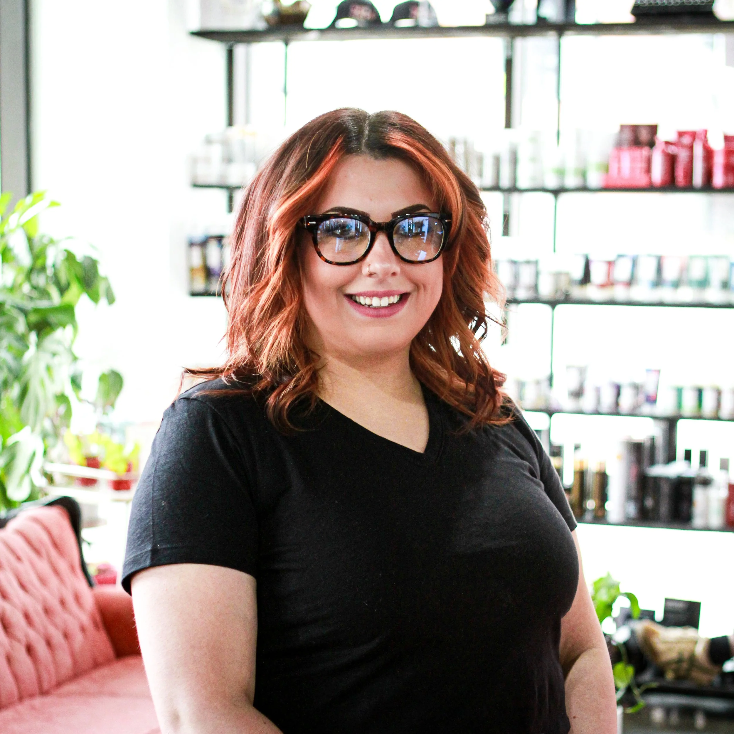 A young woman with short, wavy hair and glasses, wearing a beige ribbed sweater and a delicate necklace, sitting in a room with shelves filled with books and plants in the background.
