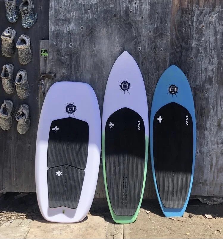 Three different surfboards leaning against a weathered wooden wall, beside a pair of rock climbing shoes.