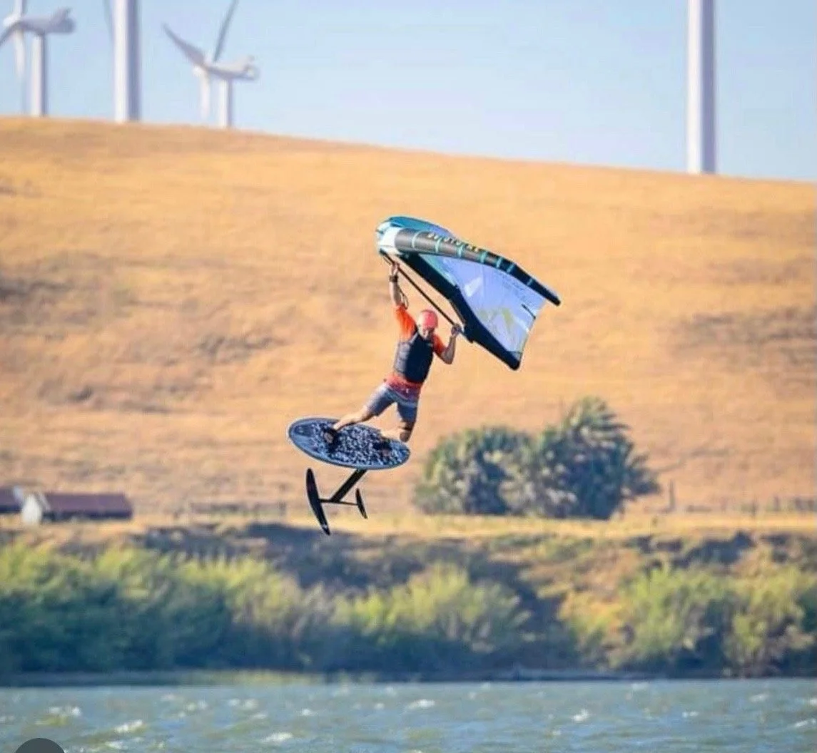 A man is wakeboarding on a body of water while flying through the air holding a large windsurfing sail, with wind turbines on the hills in the background.