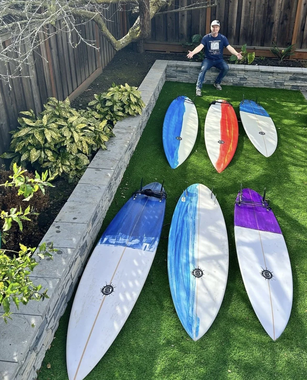 An outdoor garden area with a man sitting on a stone retaining wall, six surfboards with colorful designs placed on grass, and a wooden fence in the background.