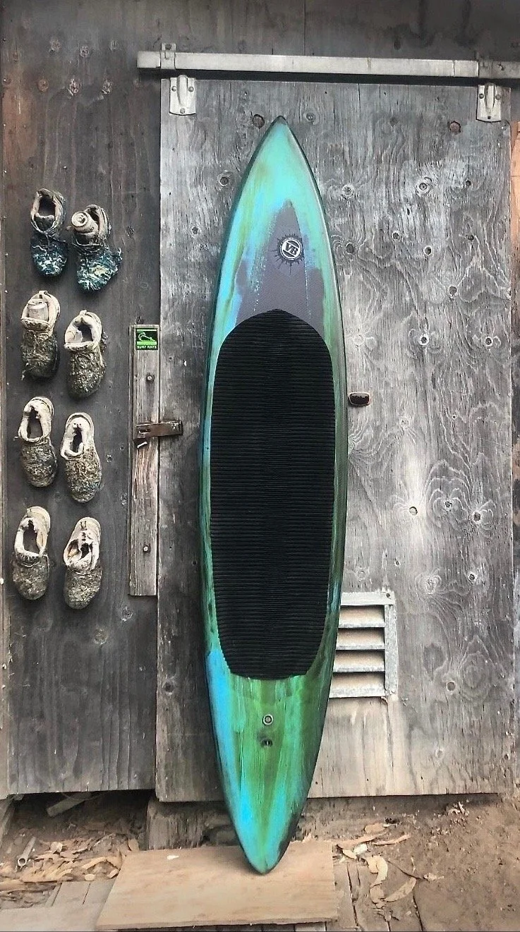 A surfboard with a black traction pad and iridescent colors leaning against a weathered wooden wall, with a pair of matching rock shoes on the left and a small vent grate near the bottom