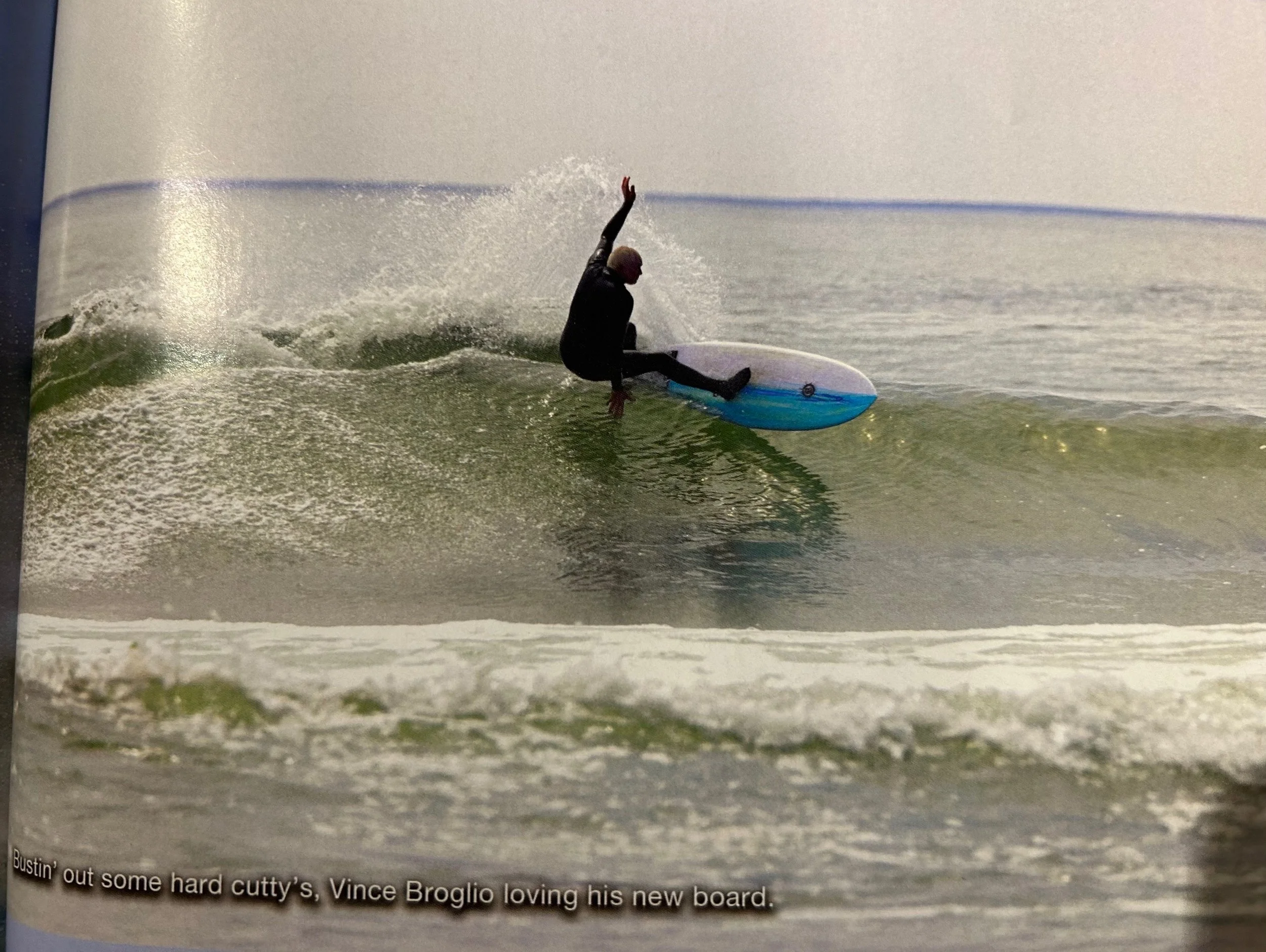 A person surfing on a wave with a blue and white surfboard, wearing a black wetsuit, raising one arm while balancing on the wave.