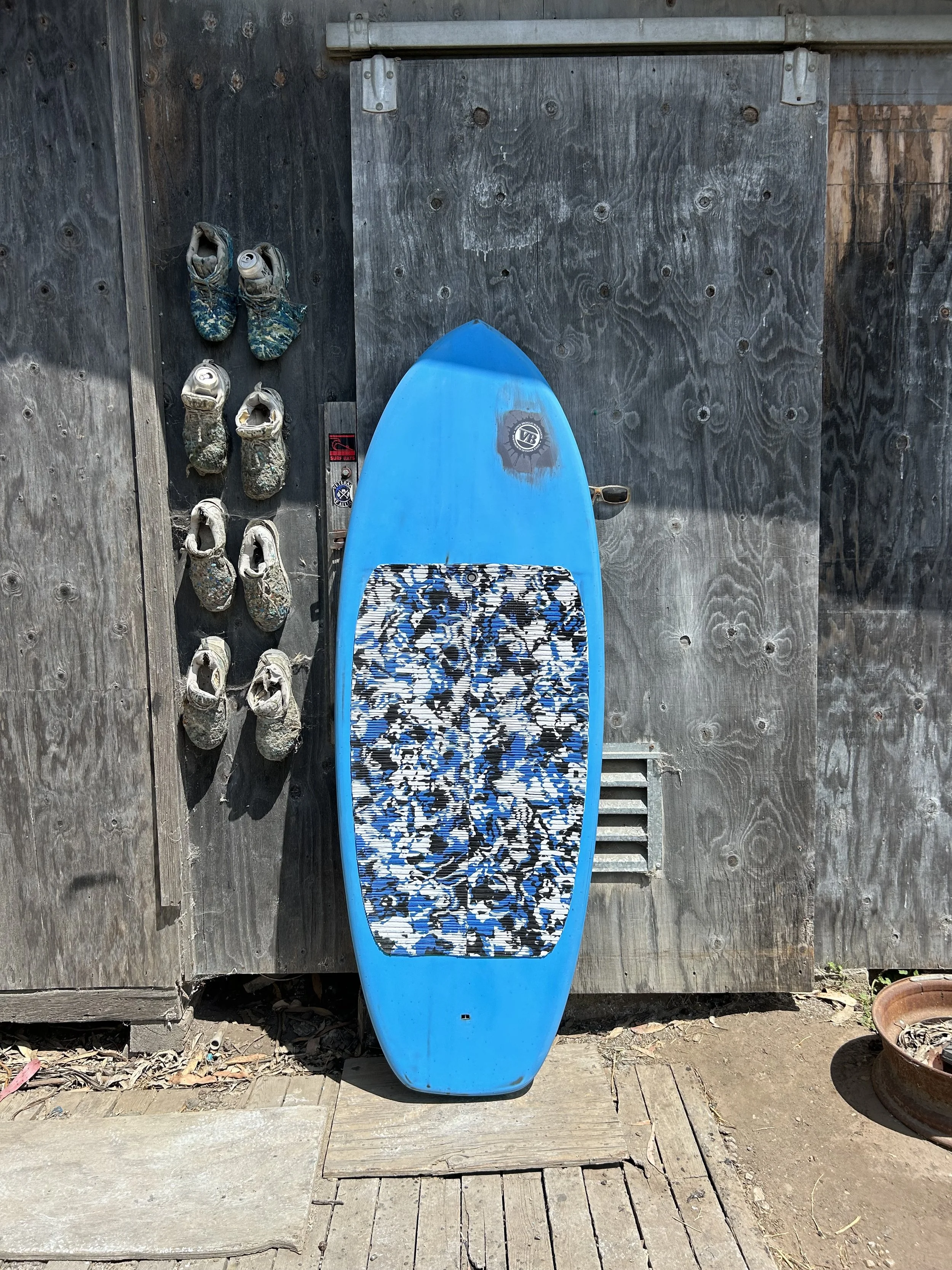 A blue and black camouflage patterned surfboard is leaning against a weathered wooden shed, with six hiking shoes hanging on the wall to the left of it, and a round plastic planter on the ground to the right.