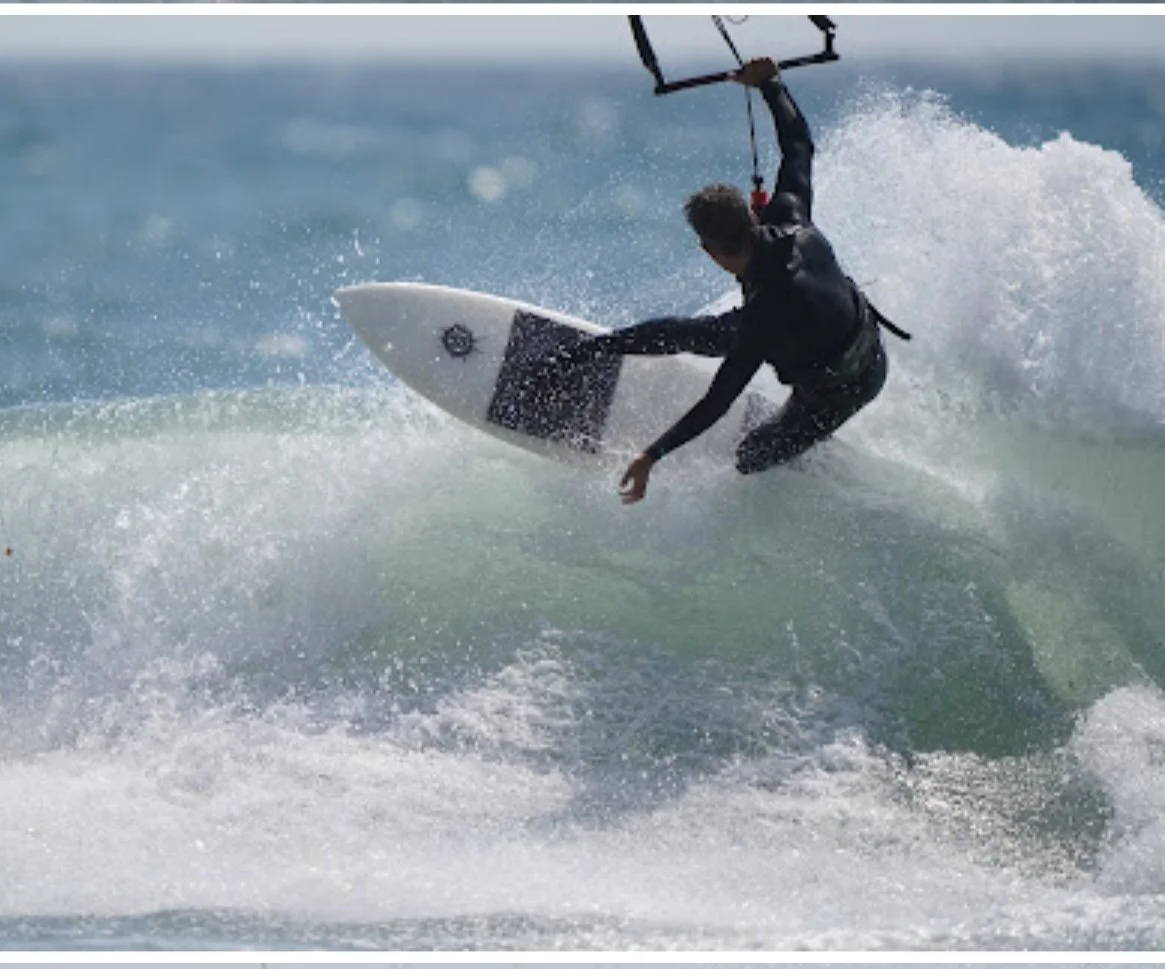 A person wearing a wetsuit riding a wave on a surfboard while holding a kite for kite surfing.