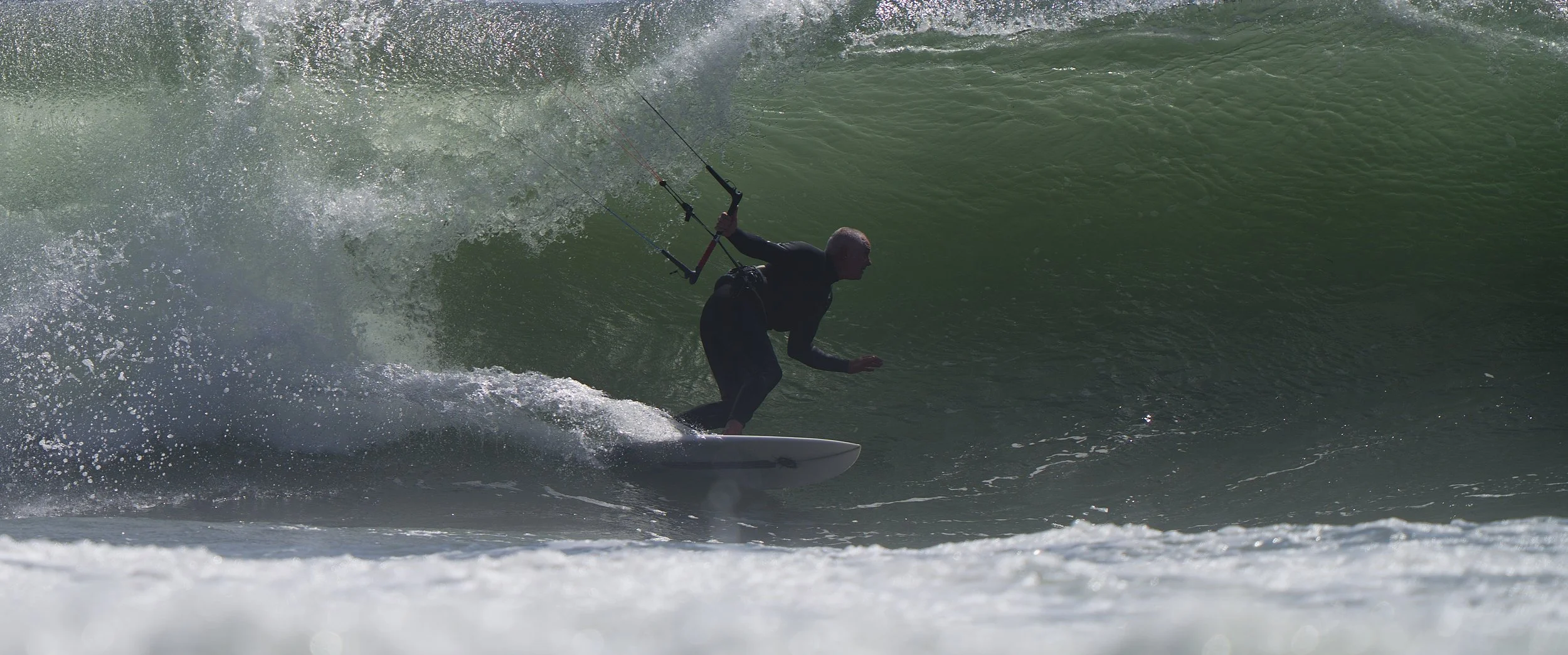 Person in black wetsuit kitesurfing on a green wave in the ocean.