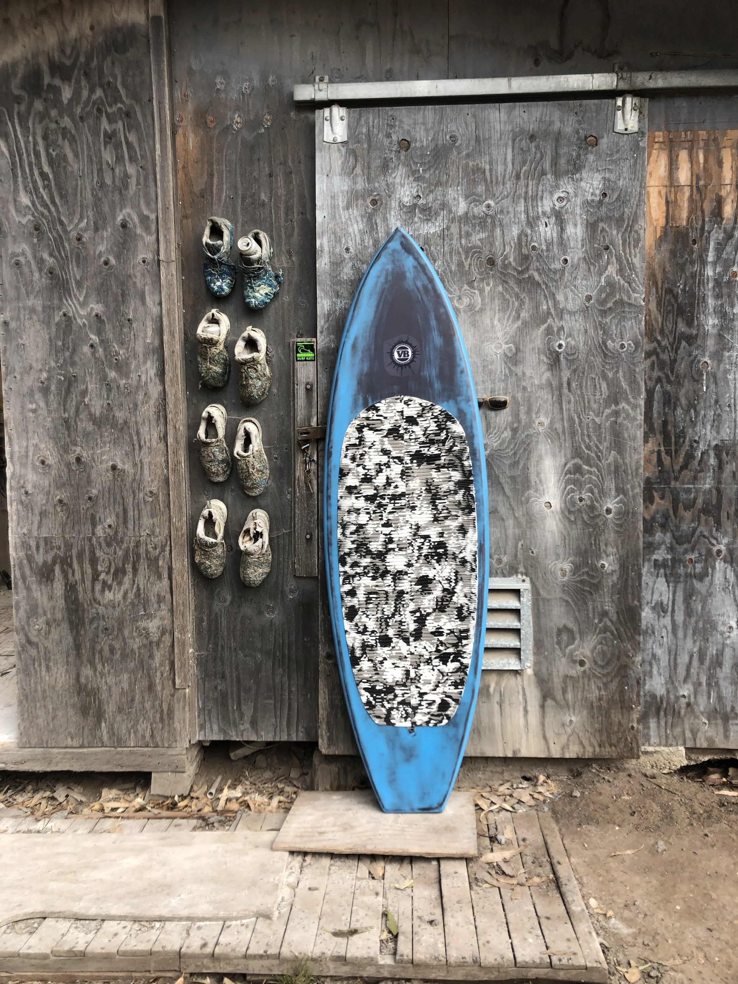 A weathered blue surfboard with a black and white patterned center rests against a rustic wooden wall, with worn shoes hanging on the left side.