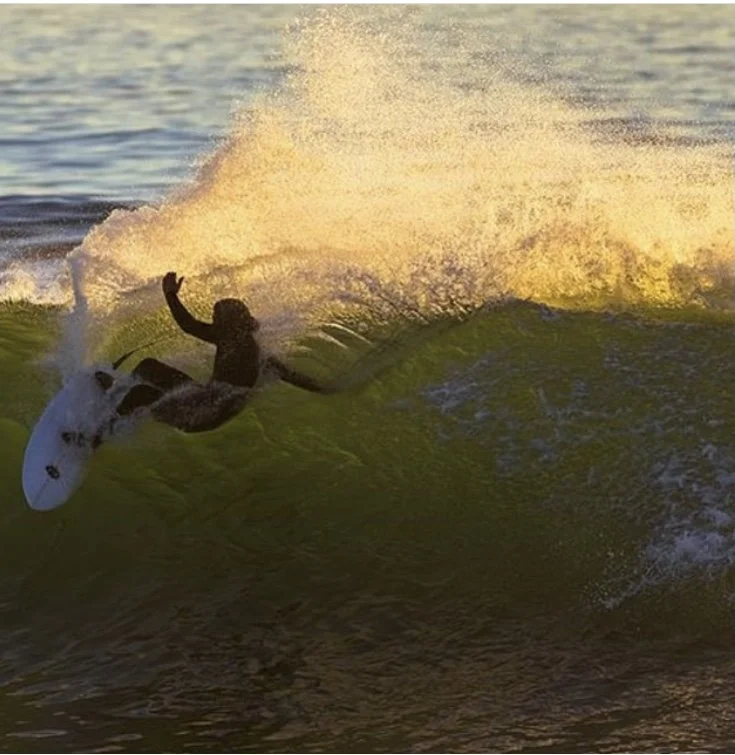 Surfer riding a wave at sunset, with spray and foam in the air.