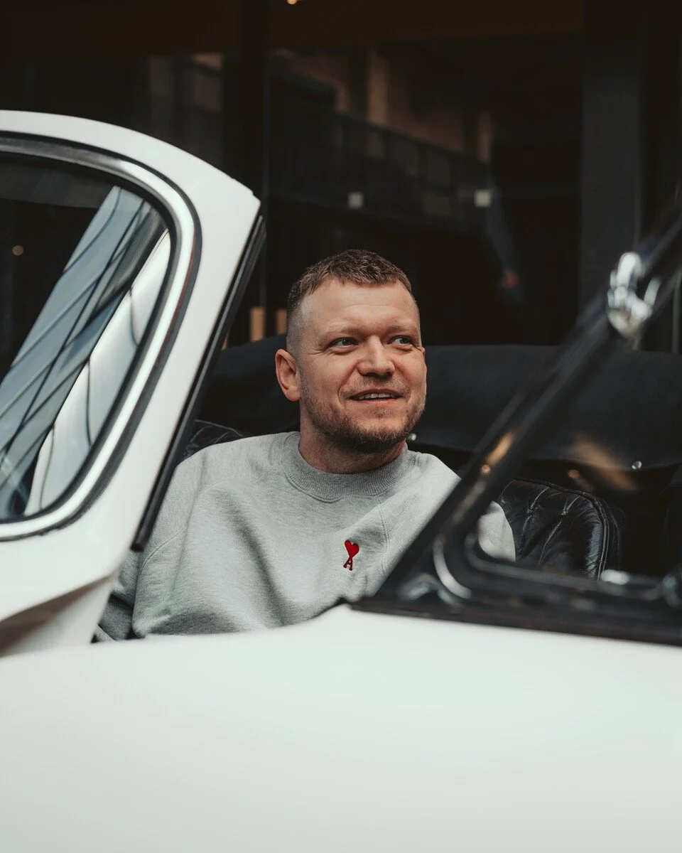 Ein Mann sitzt in einem weißen Cabrio, trägt ein graues Sweatshirt mit einem kleinen roten Herzen auf der linken Seite der Brust. Er lächelt leicht und schaut nach rechts.