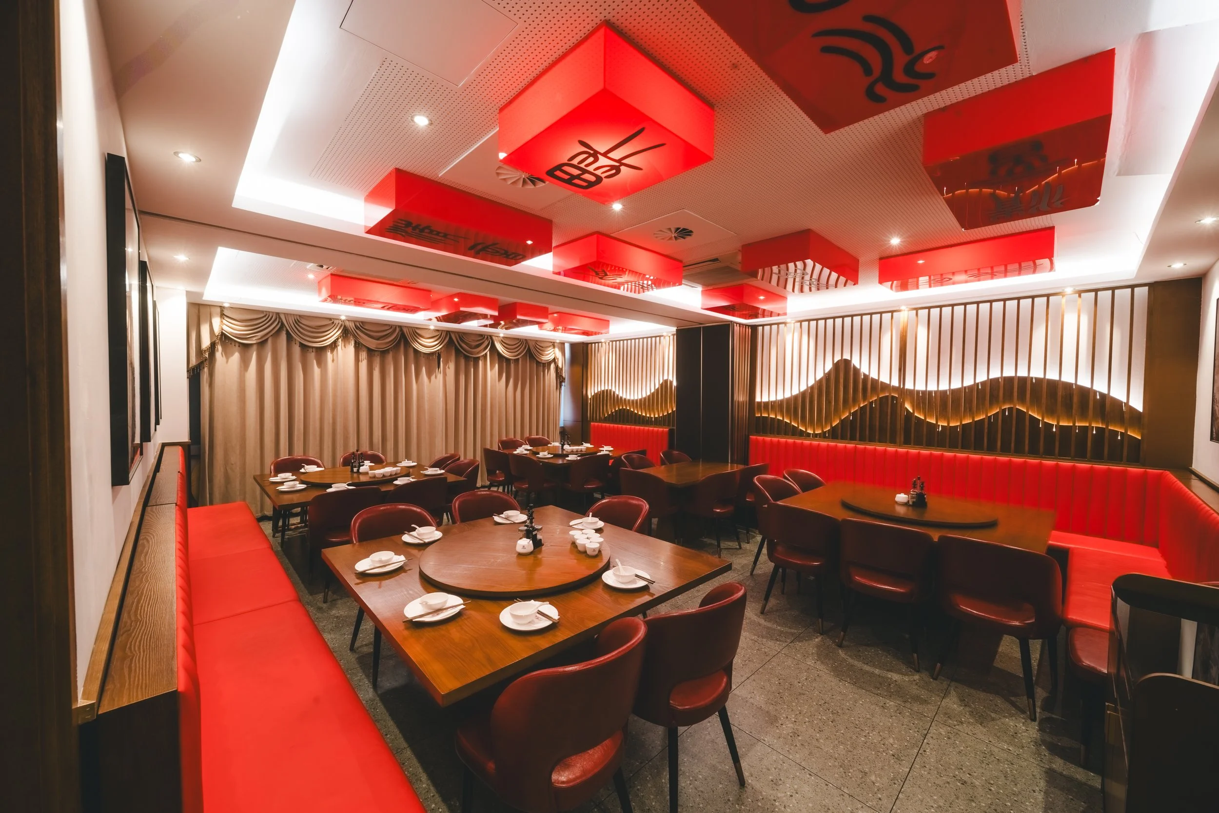 Ein elegante japanische Restaurant mit roten und schwarzen Dekorelementen an der Decke und einem hohen, geraden Holzverteil mit roten gepolsterten Bänken und Stühlen am Esstisch. Die Tische sind mit Tellern und Besteck vorbereitet.