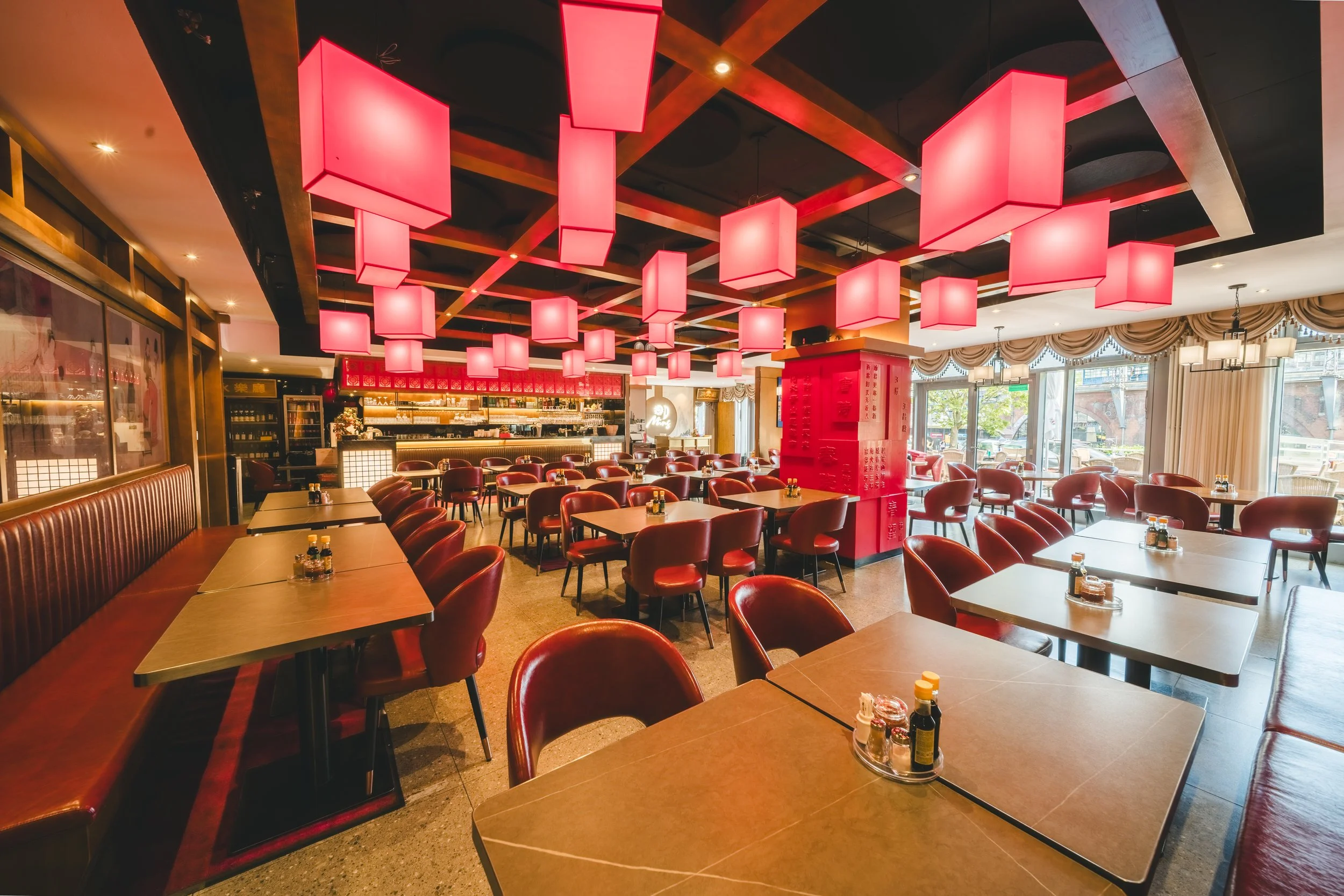 Innenansicht eines Restaurants mit roten Stühlen, dunklen Tischen und einer Decke mit roten Lampen in rechteckiger Form.