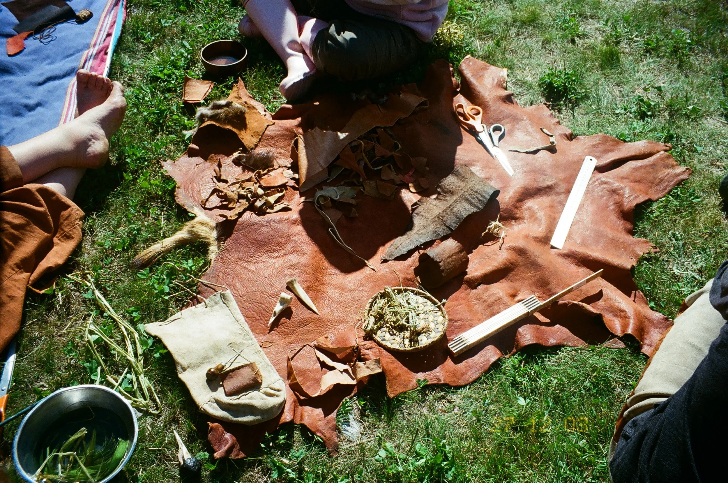 A person sitting on grass next to a leather hide with various tools and objects spread out on it, including scissors, a ruler, and a small woven basket, with a cup of tea nearby on a tent or blanket.
