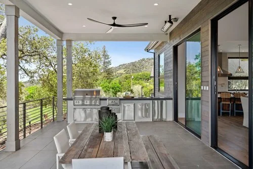 This photo captures a beautiful outdoor patio space that is covered by a roof. The patio is furnished with an elegant outdoor dining set, featuring a spacious table and comfortable chairs. Adjacent to the dining area, there is an outdoor kitchen, com