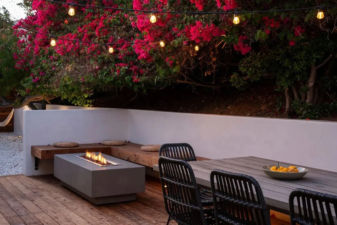 Luxury Malibu outdoor living space with a modern fire pit, custom wood bench, outdoor dining area, landscape lighting, and blooming bougainvillea.