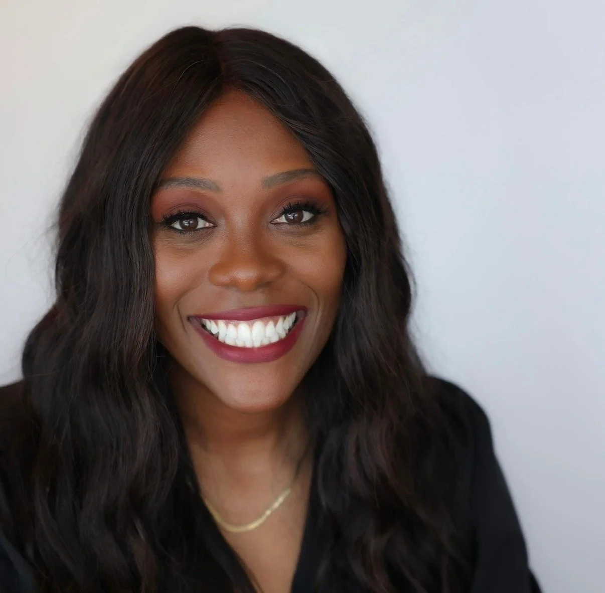 Close-up of a smiling Black woman with long wavy black hair, wearing a black top and a gold necklace, against a plain light background.
