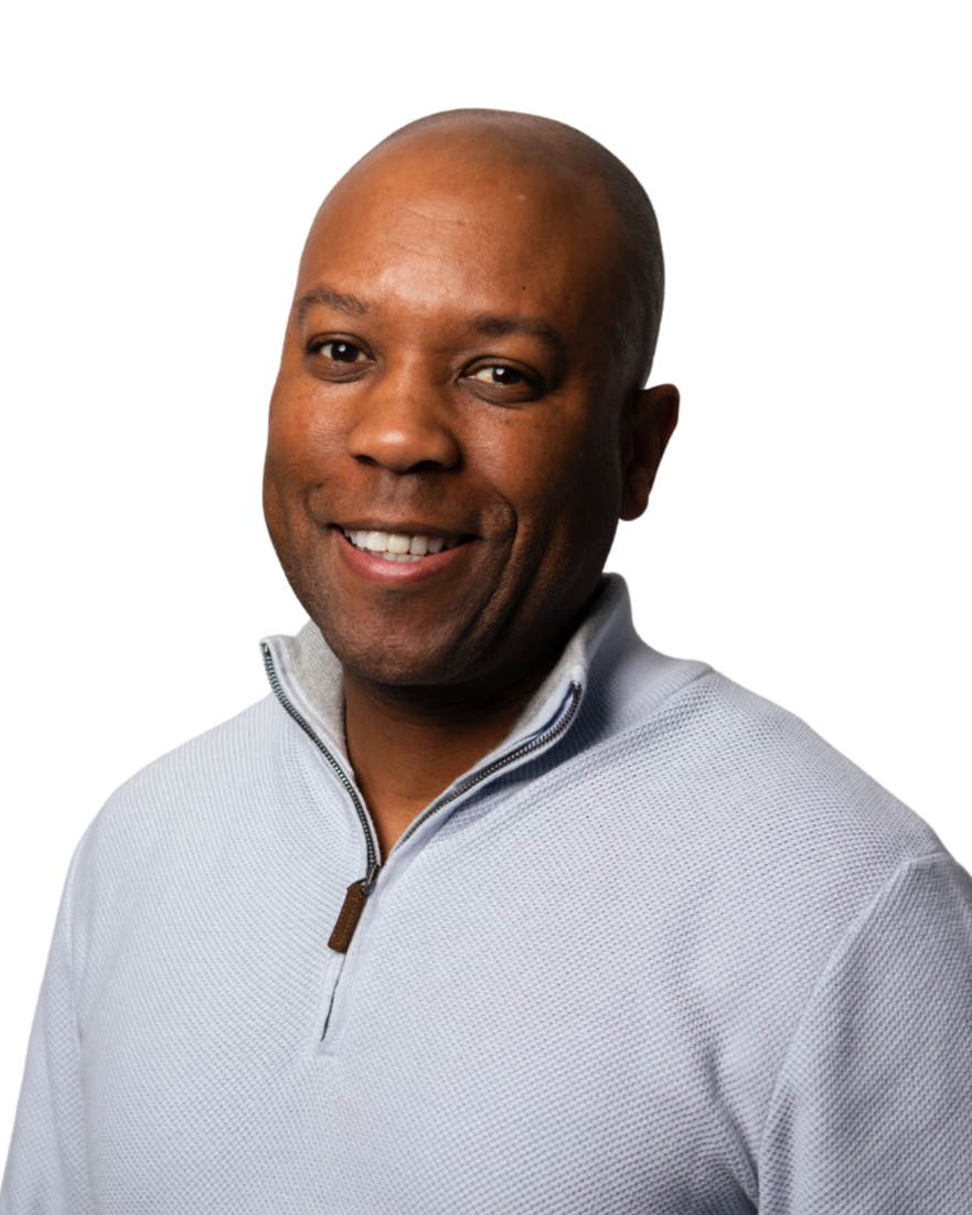 A smiling African American man with a shaved head wearing a light gray quarter-zip sweater against a white background.