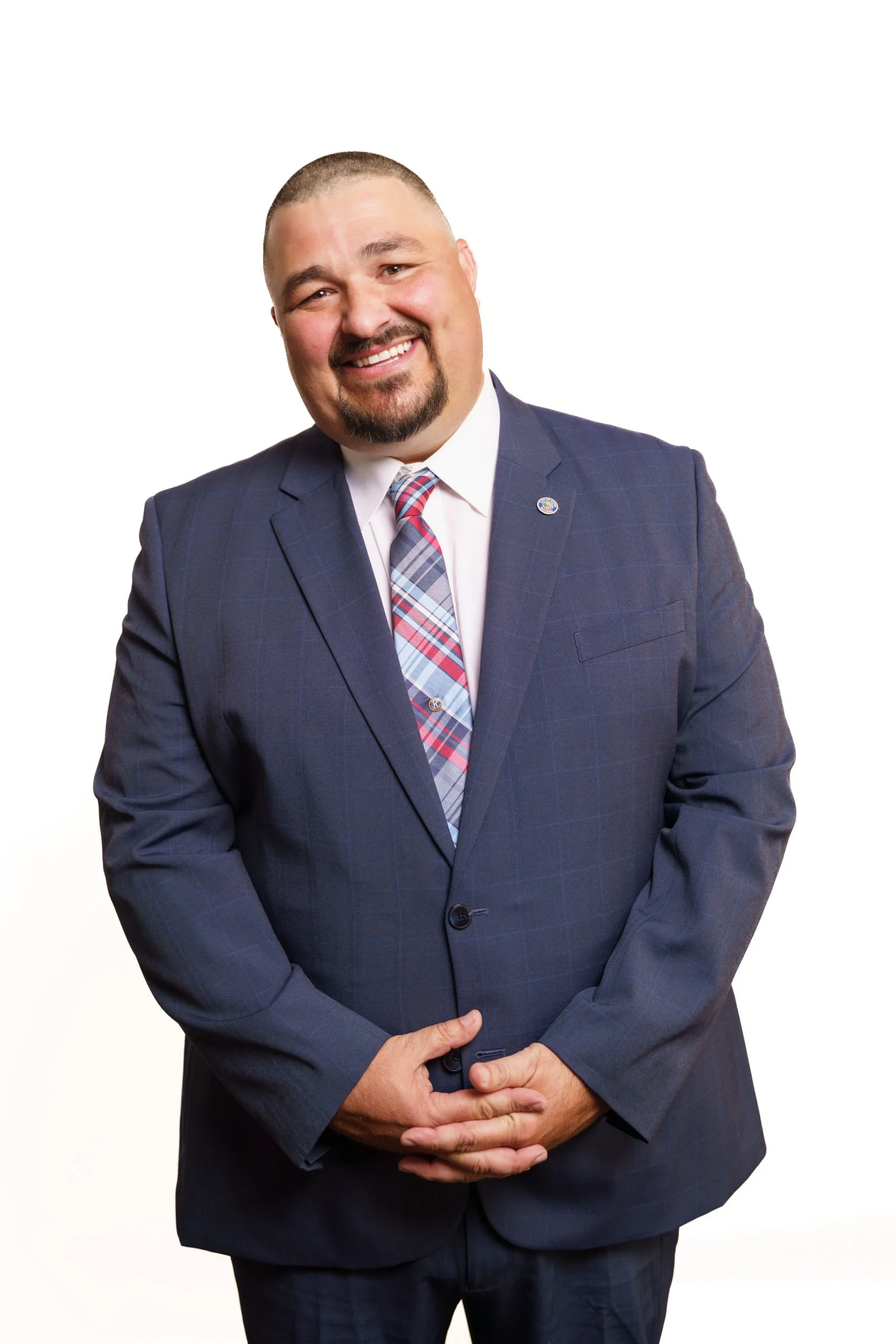 A man in a dark blue suit, white shirt, and patterned tie, smiling with his hands clasped in front, standing against a plain white background.