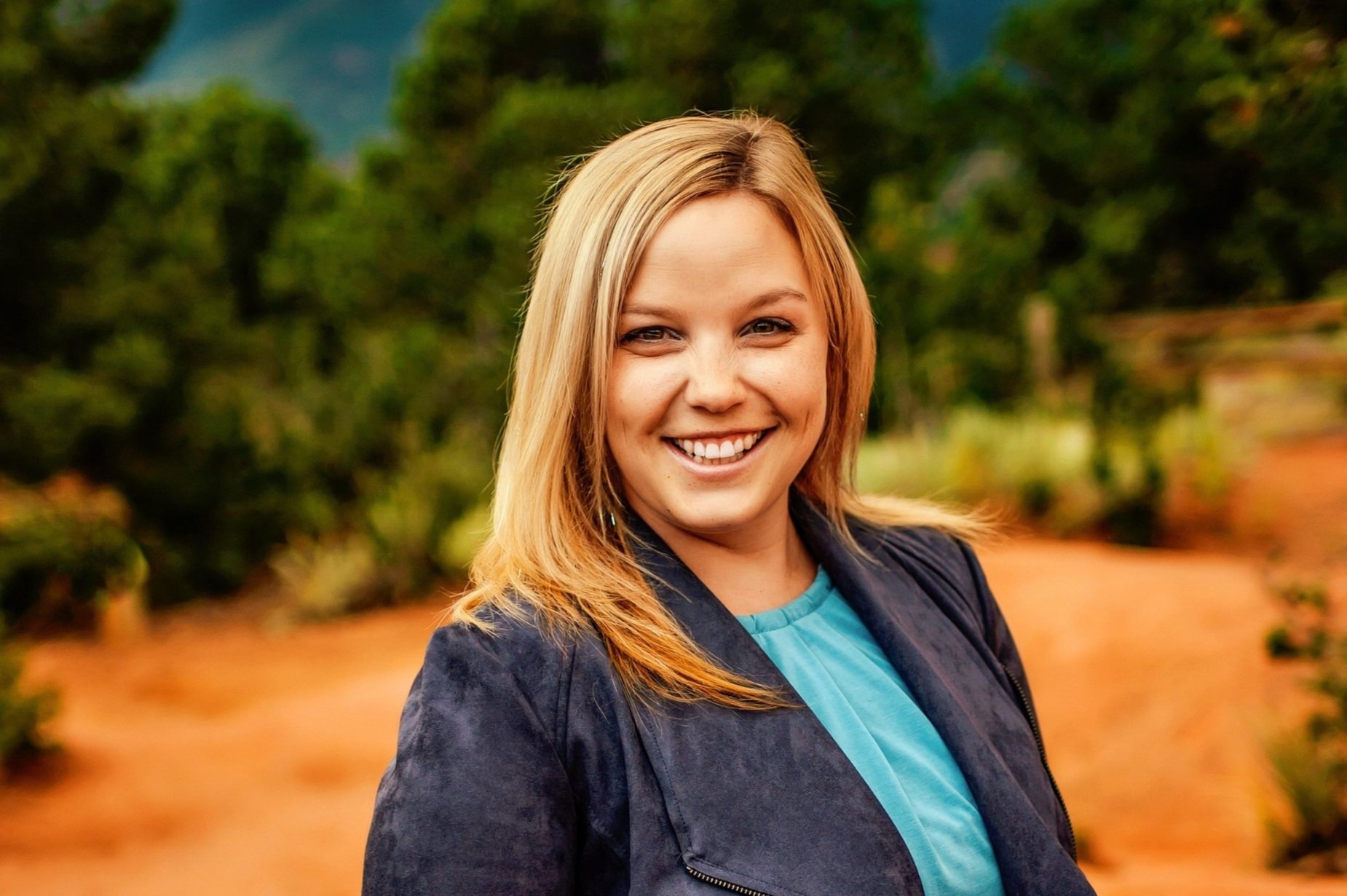 Smiling woman with blonde hair in a blue jacket standing outdoors with trees and mountains in the background.