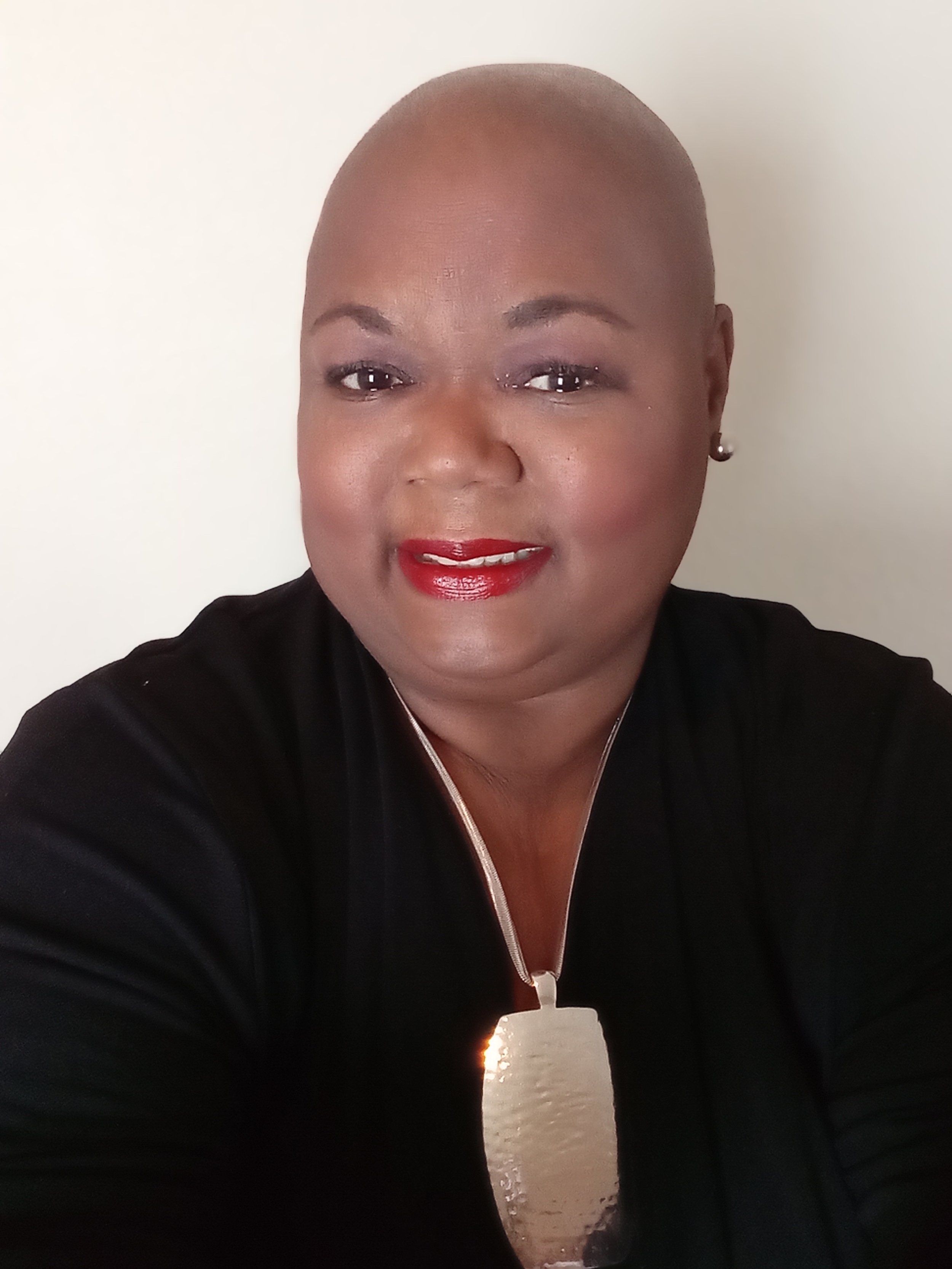 A person with a bald head, wearing a black outfit and a gold necklace, with red lipstick and pearl earrings, smiling against a light background.