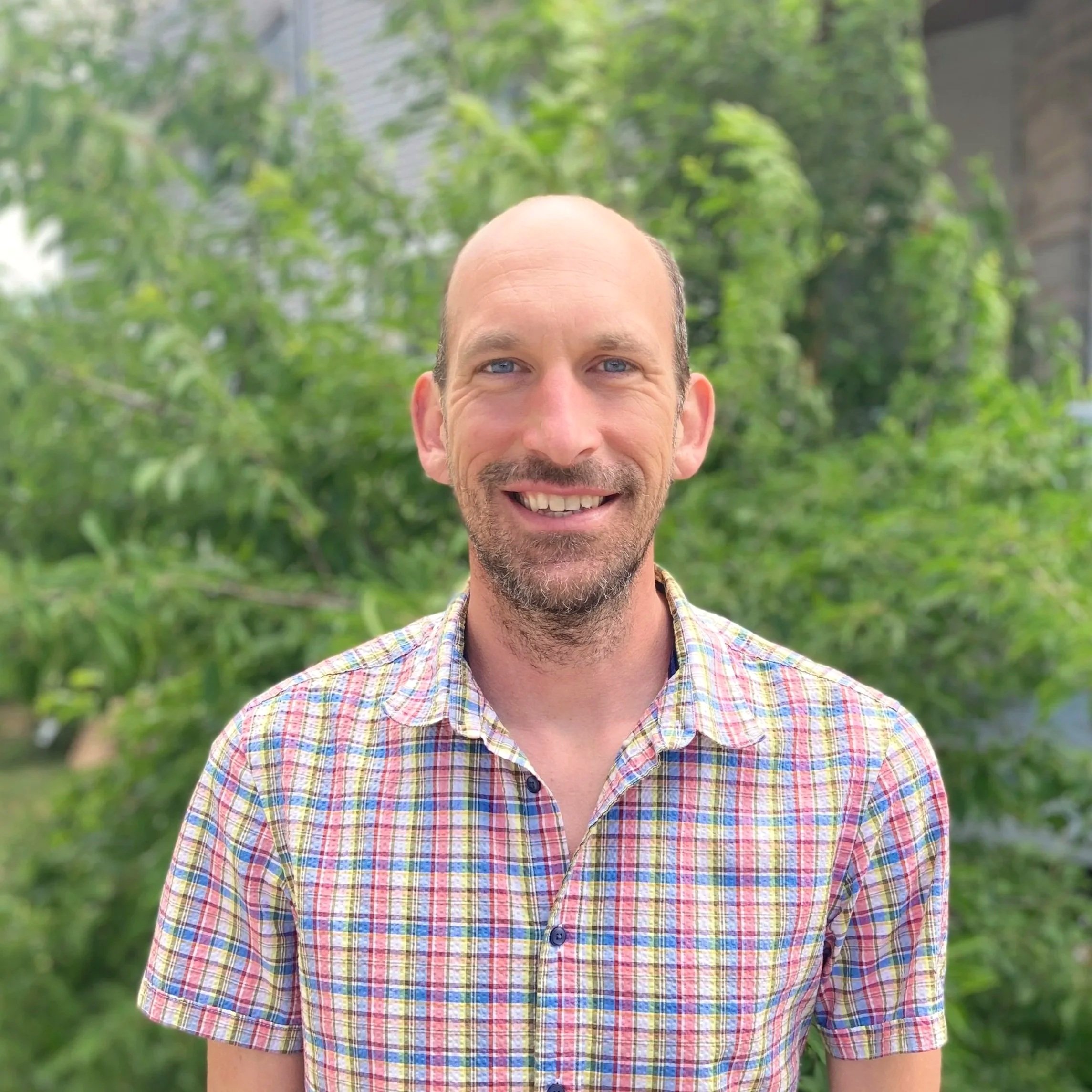White balding man with a beard in a plaid shirt smiling 