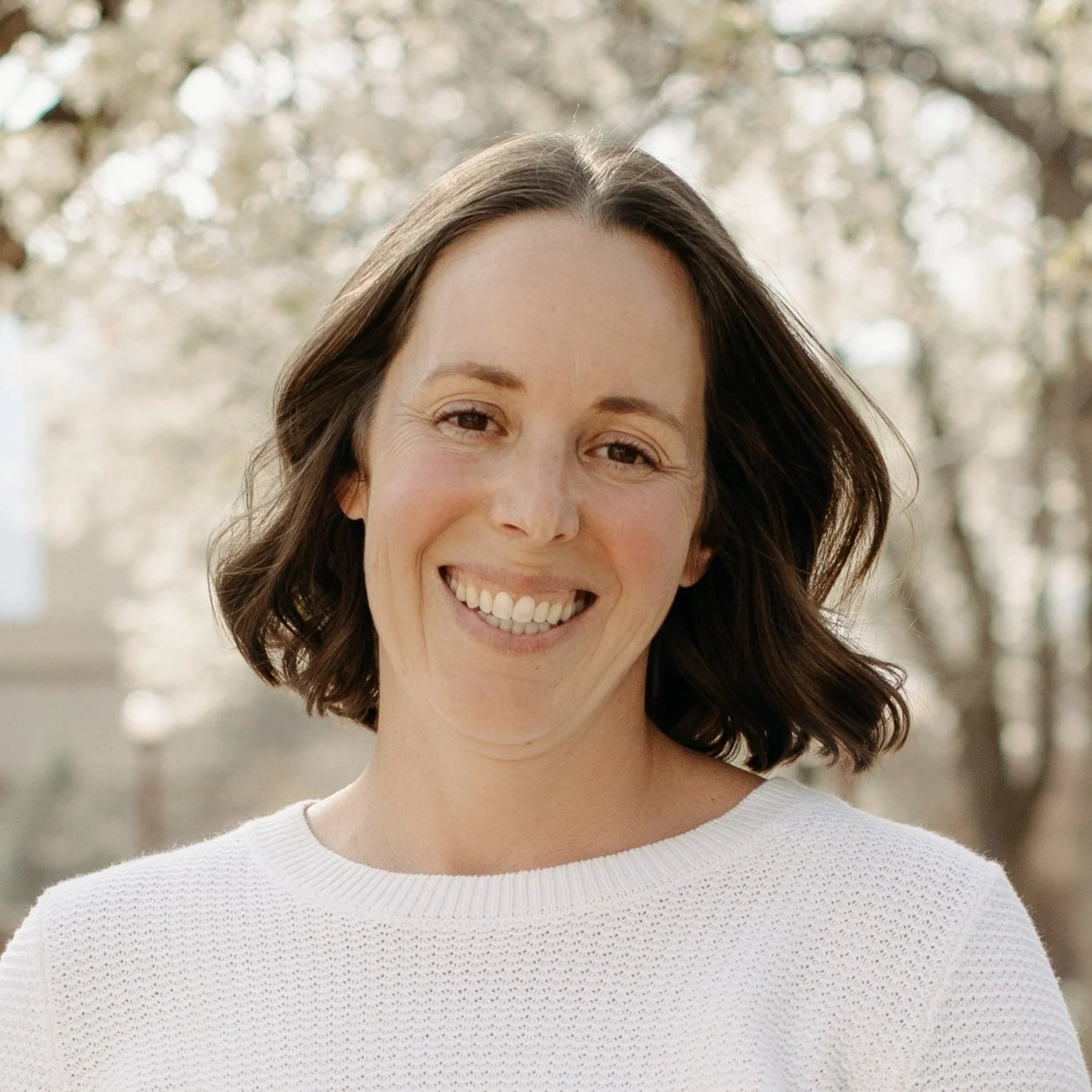 Smiling woman with shoulder-length brown hair in a white sweater, outdoor setting.