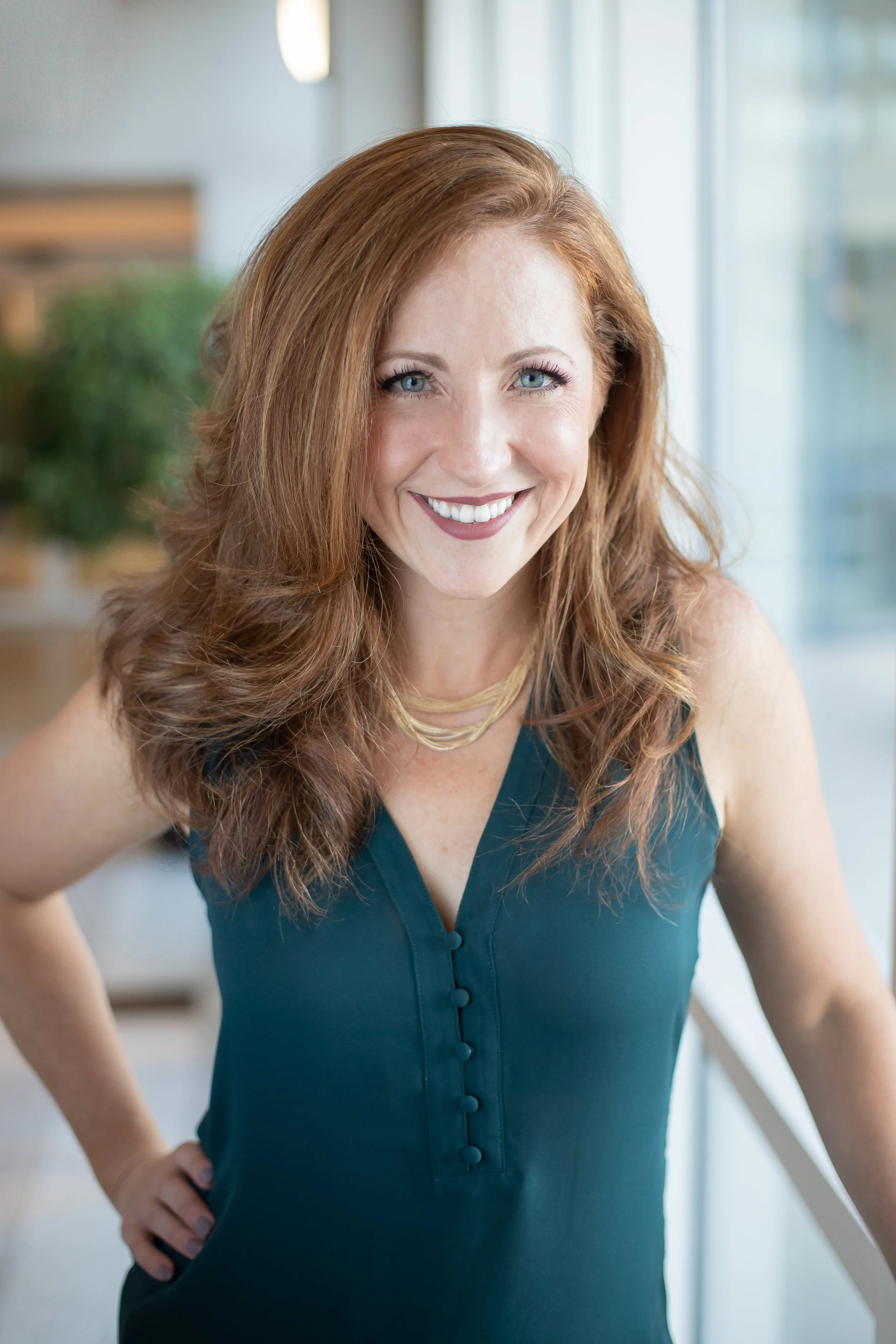Smiling woman with red hair and teal sleeveless top, standing indoors by a window.