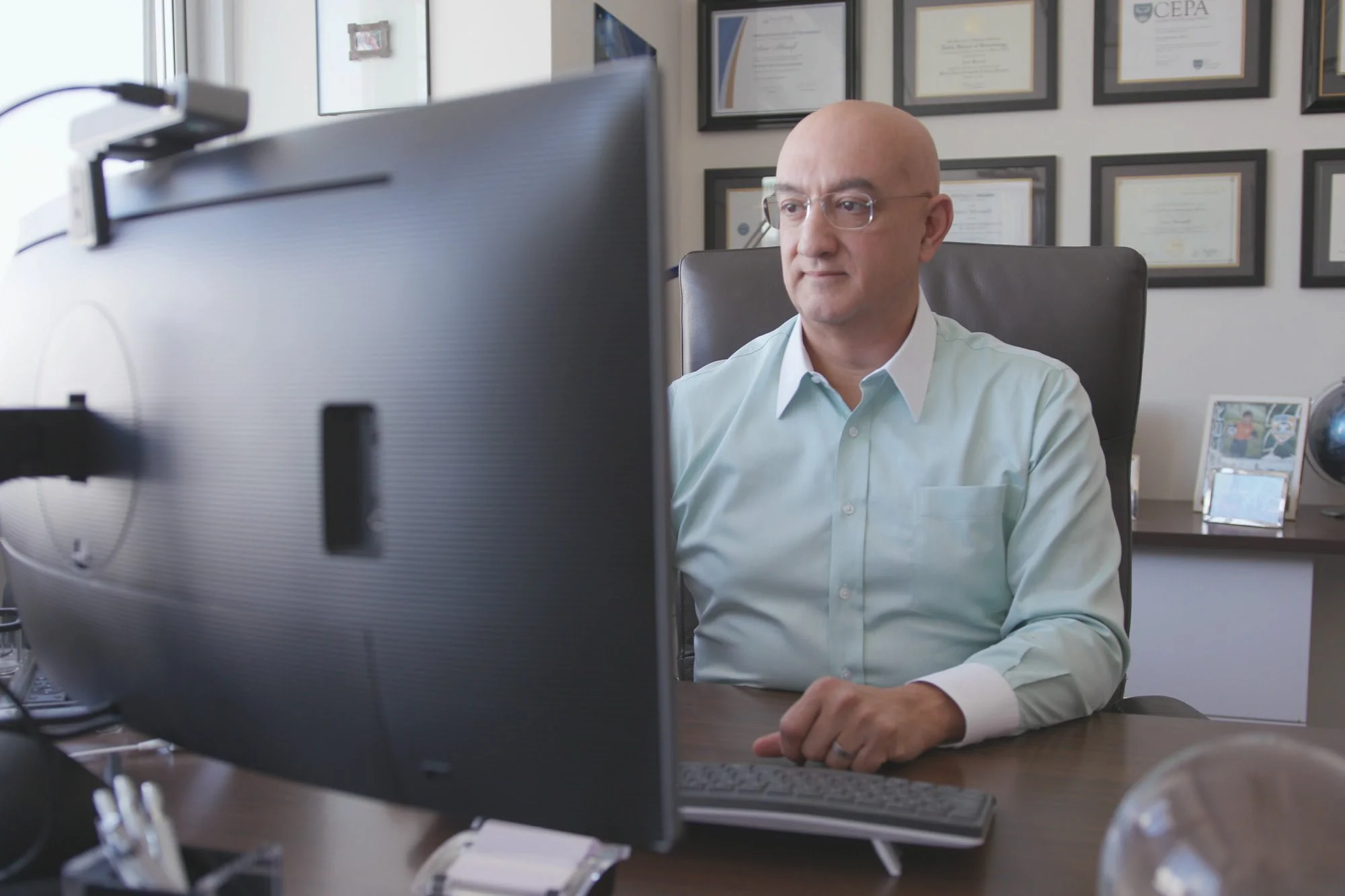 A man wearing glasses and a light green dress shirt sits at a desk, looking at a computer monitor in an office with framed certificates on the wall behind him.