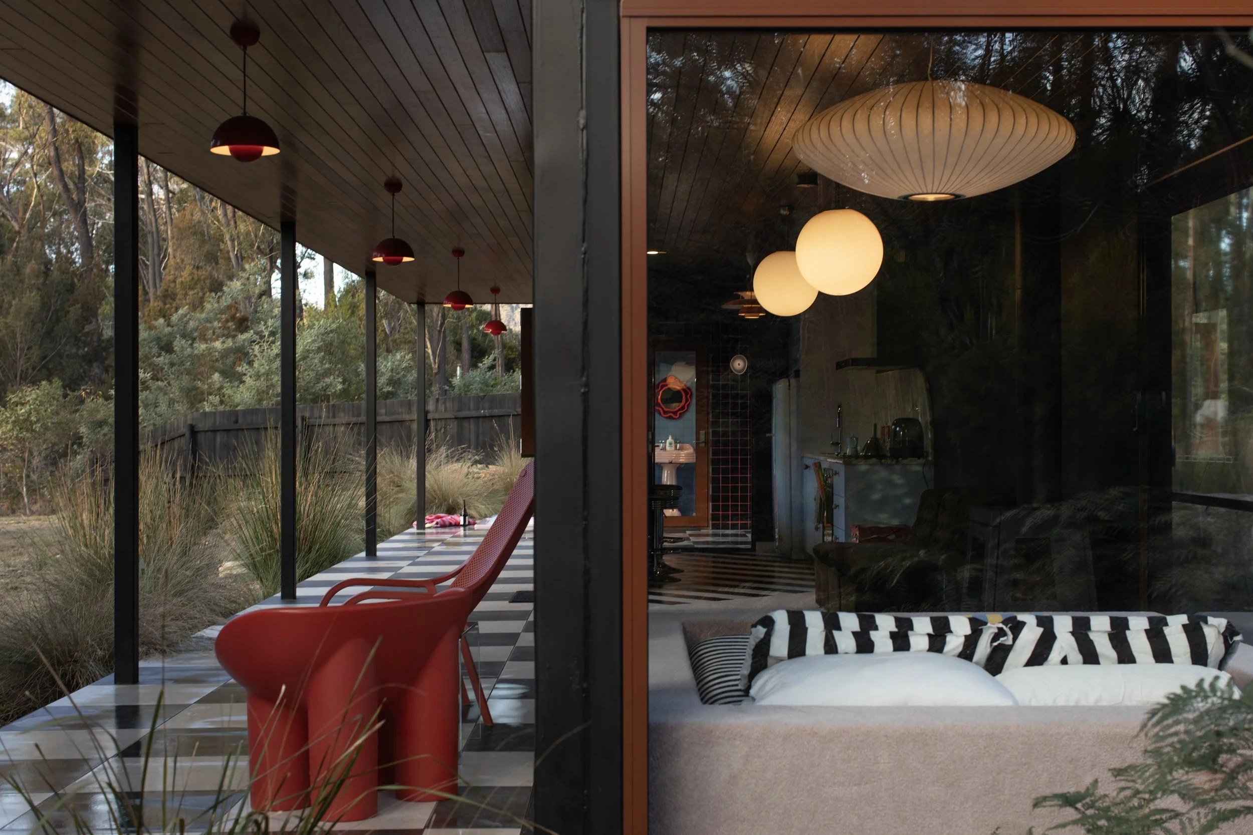 View through a large glass window showing an indoor room with a ceiling lantern, two hanging spherical lamps, and a wooden ceiling. Outside, a covered patio with red chairs and a garden with trees and grass are visible.