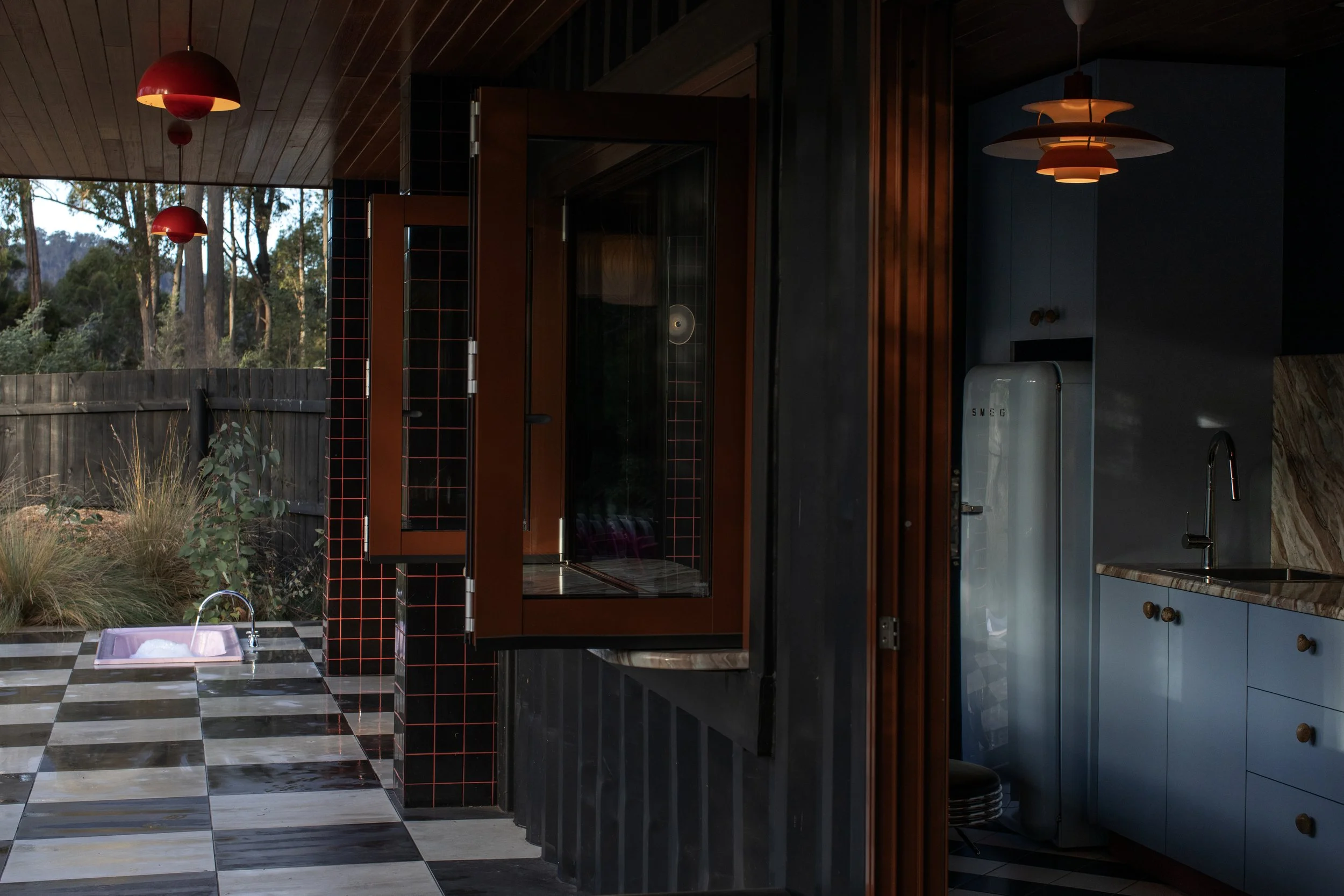 Interior view of a modern kitchen with black and wooden cabinetry, checkered black and white floor tiles, a pink sink, and modern lighting fixtures. Outside window shows trees and a fence.