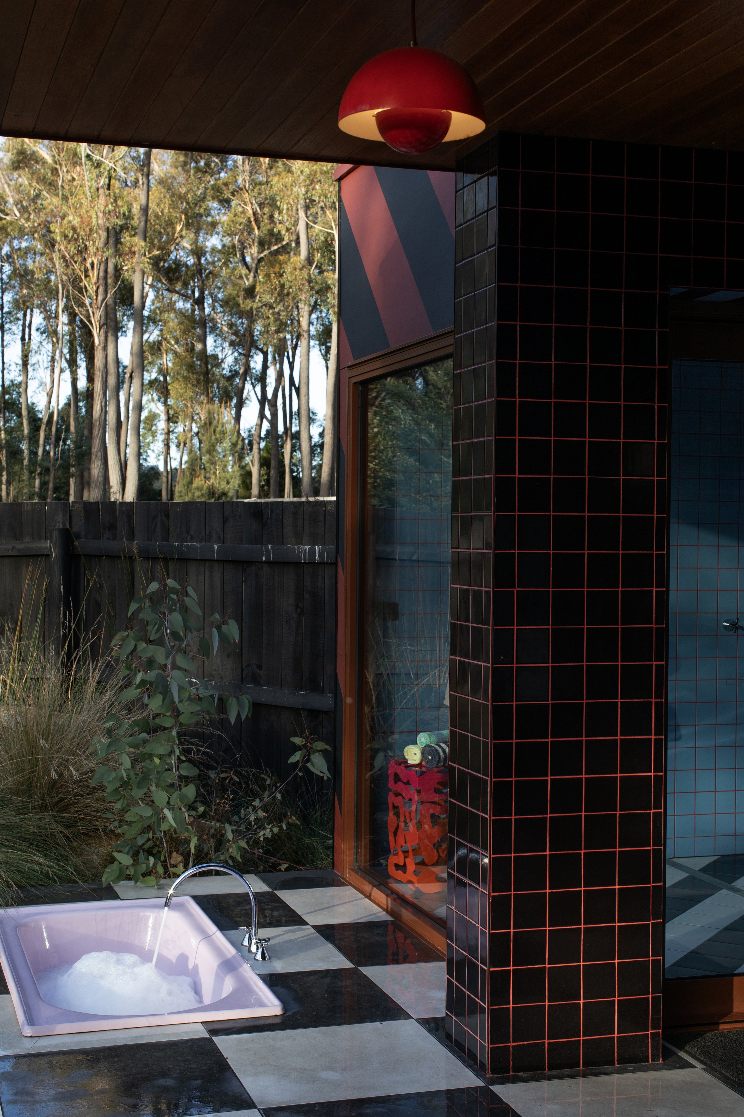Indoor pool area with black and white checkered tile flooring, a pink lounge chair with bubbles, green plants, a black tiled wall with red grout, and a window reflecting an outdoor wooded area.