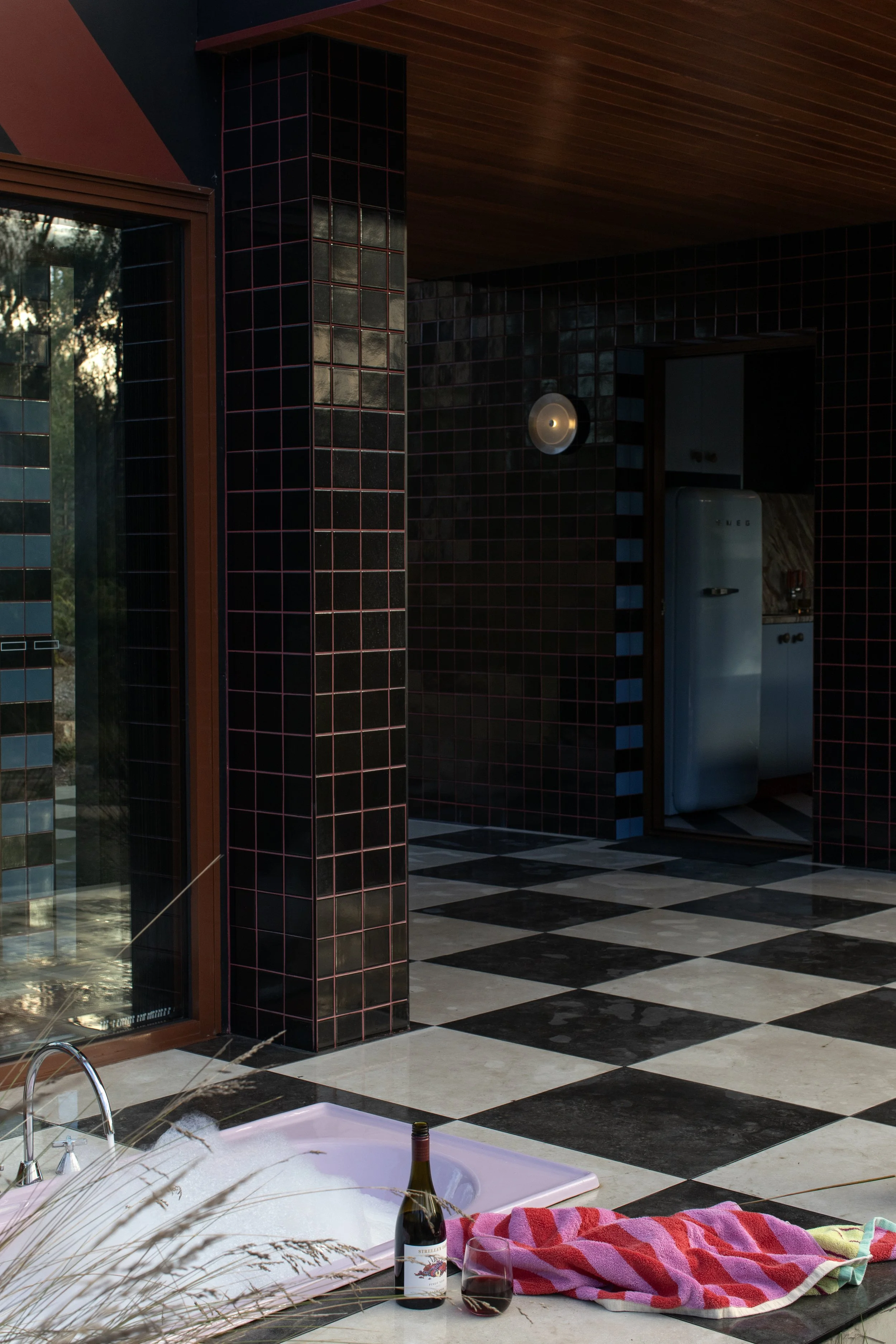 Indoor scene with a hot tub surrounded by a black and white checkered floor, a pink towel, a bottle of wine, a glass of red wine, and an open book or magazine. The background shows black tiled walls with a partial view of a refrigerator and a doorway